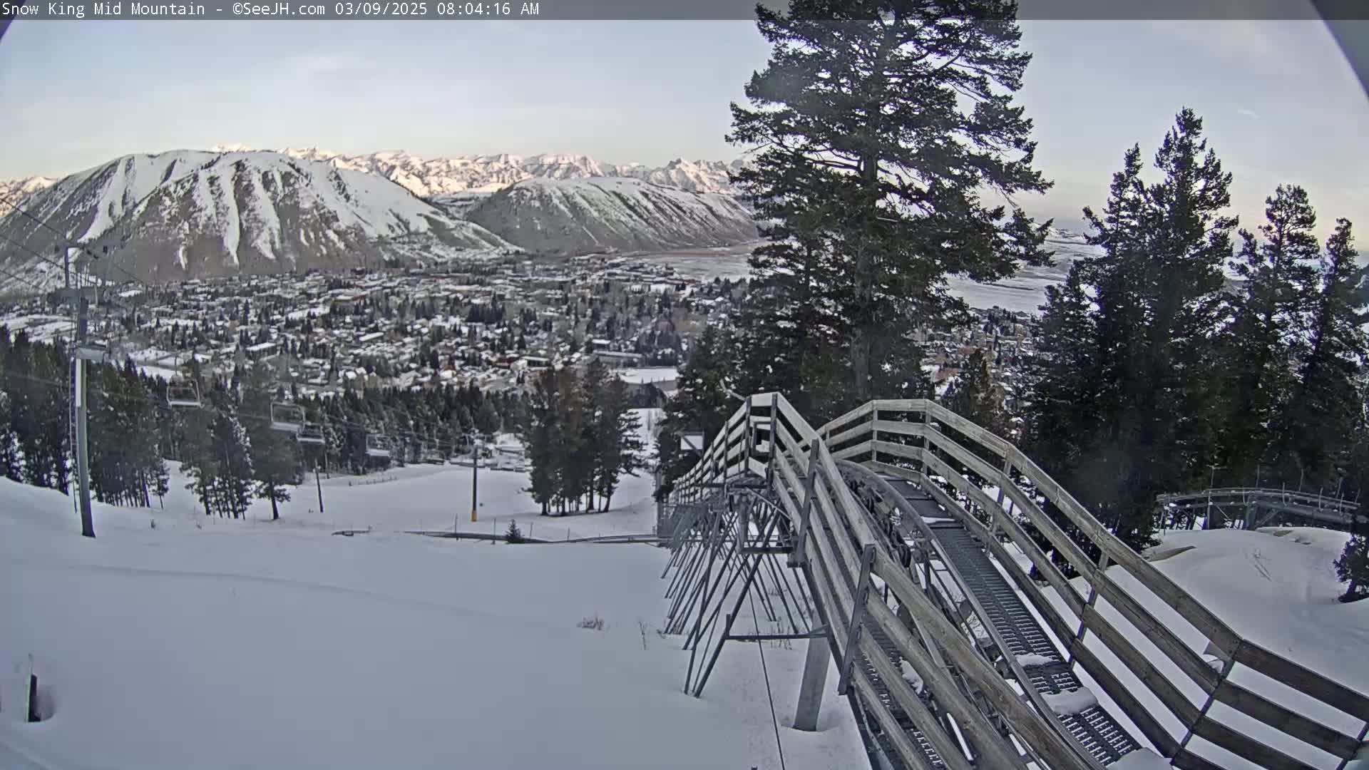 A snow-covered mountain town is visible in the distance, viewed from a snowy mountainside with a wooden alpine coaster in the foreground, under a mostly clear sky.