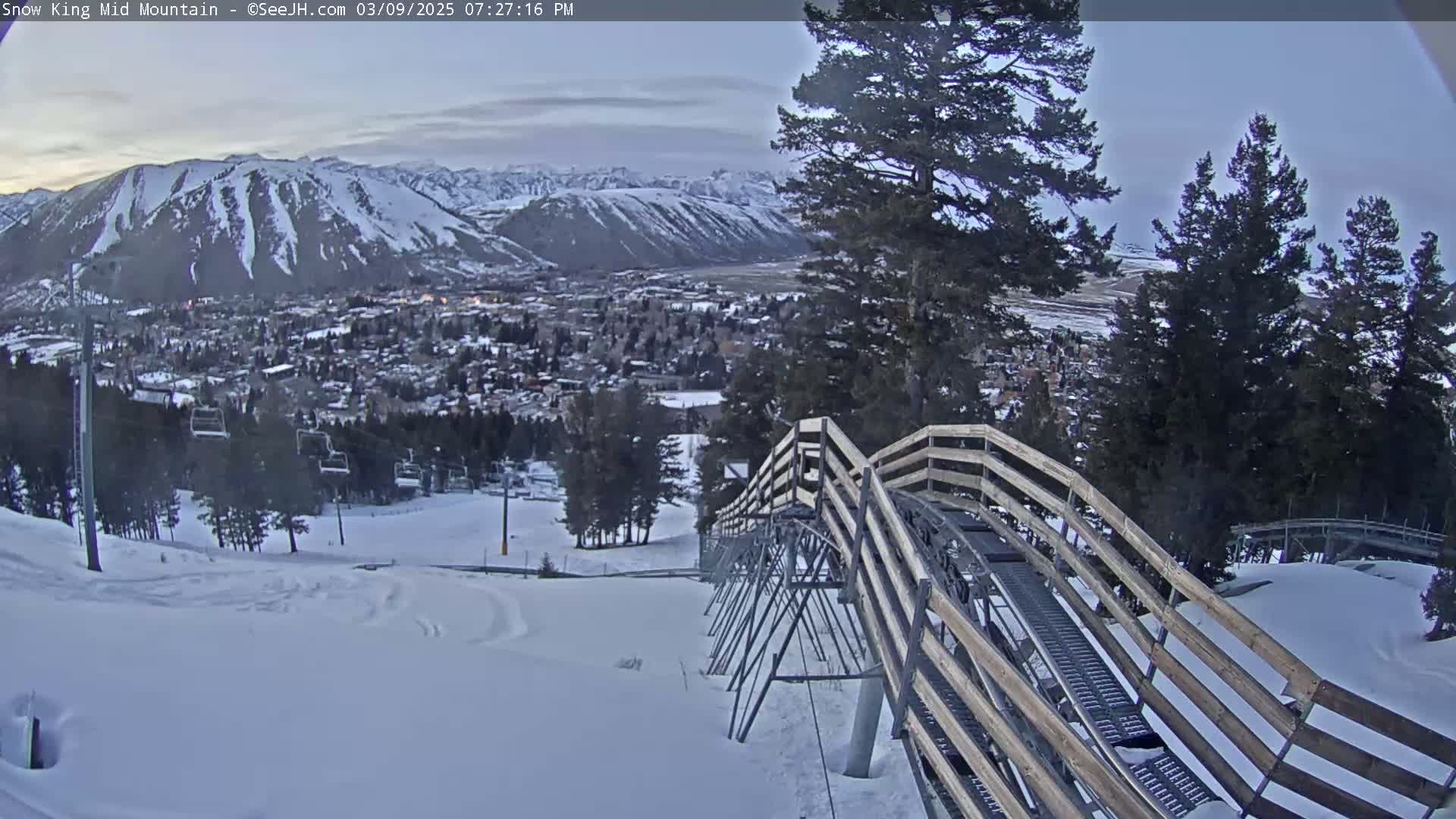 A snow-covered mountain landscape is viewed from a snow-covered alpine coaster, with a town visible in the distance under a twilight sky.