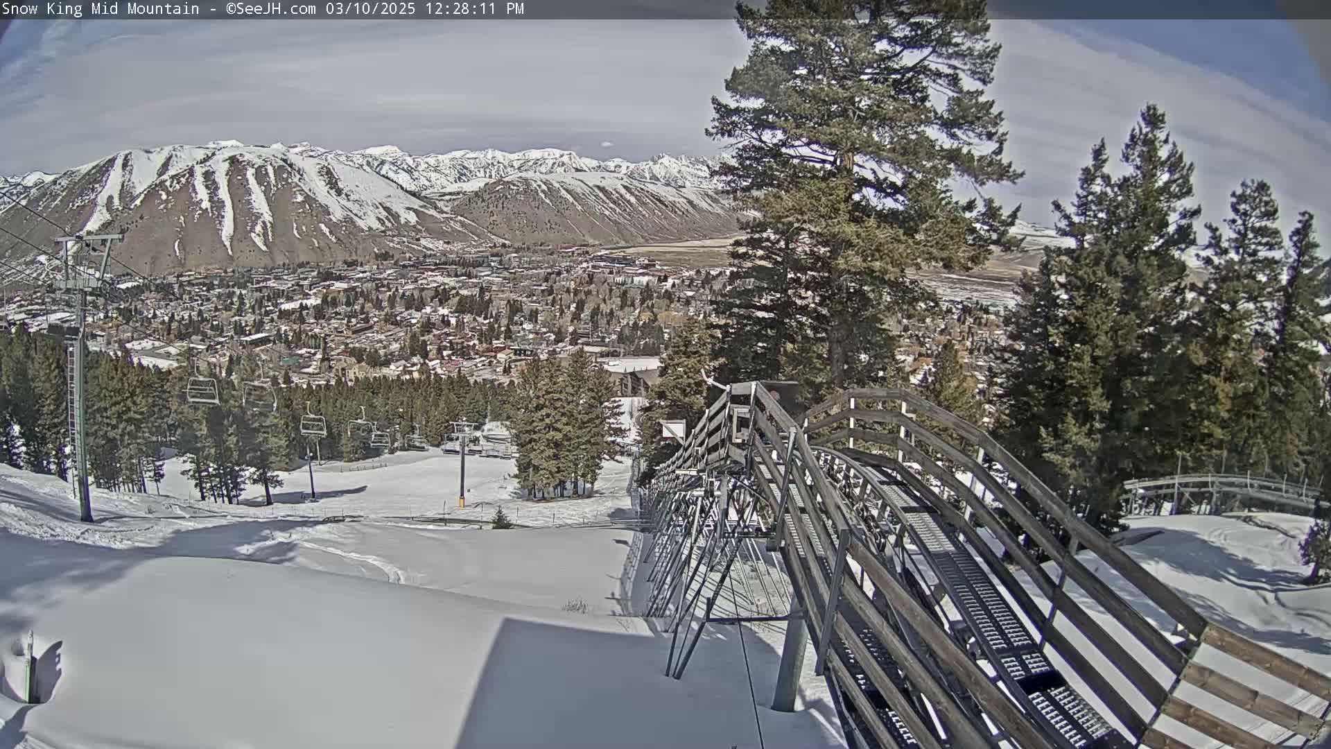 A snow-covered mountainside with a ski lift and a town in the valley below on a partly sunny day.