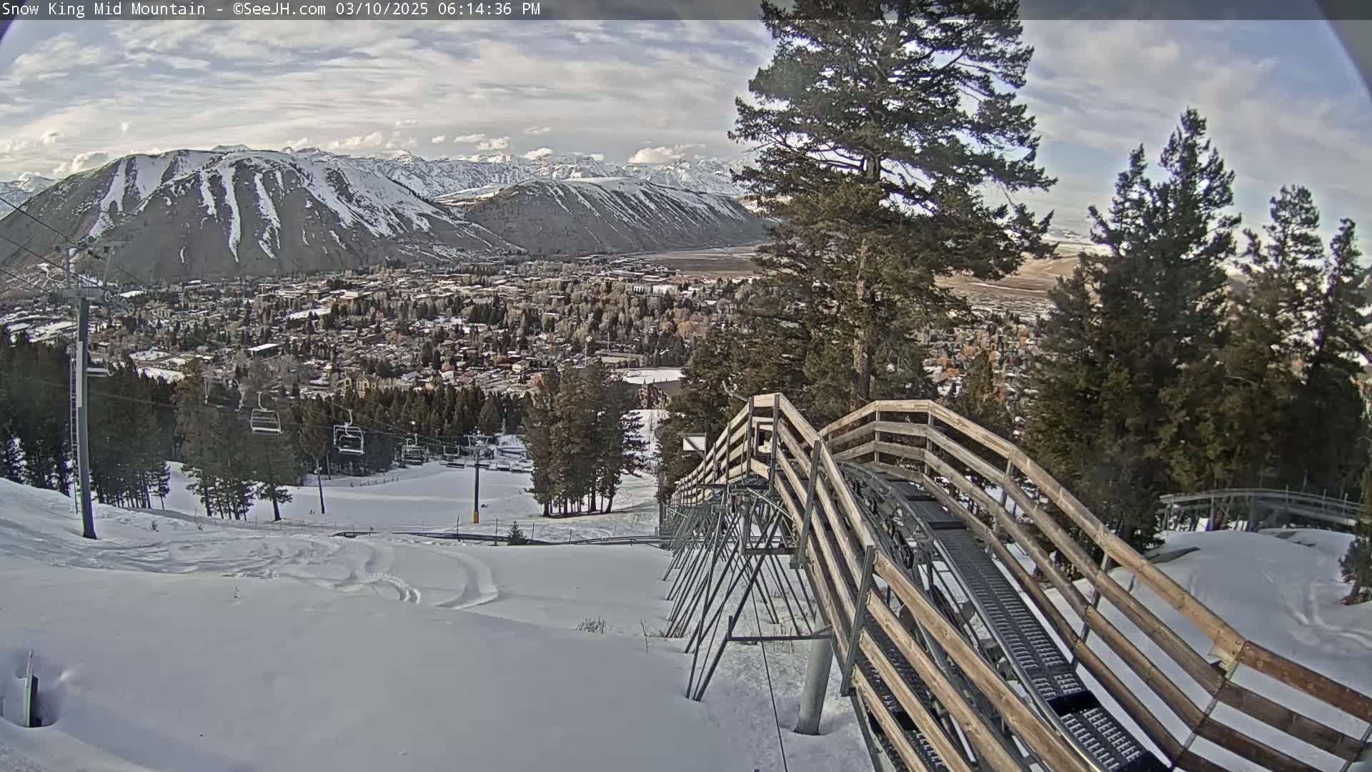 A snow-covered mountain slope with a wooden alpine coaster overlooks a town nestled in a valley under a partly cloudy sky.