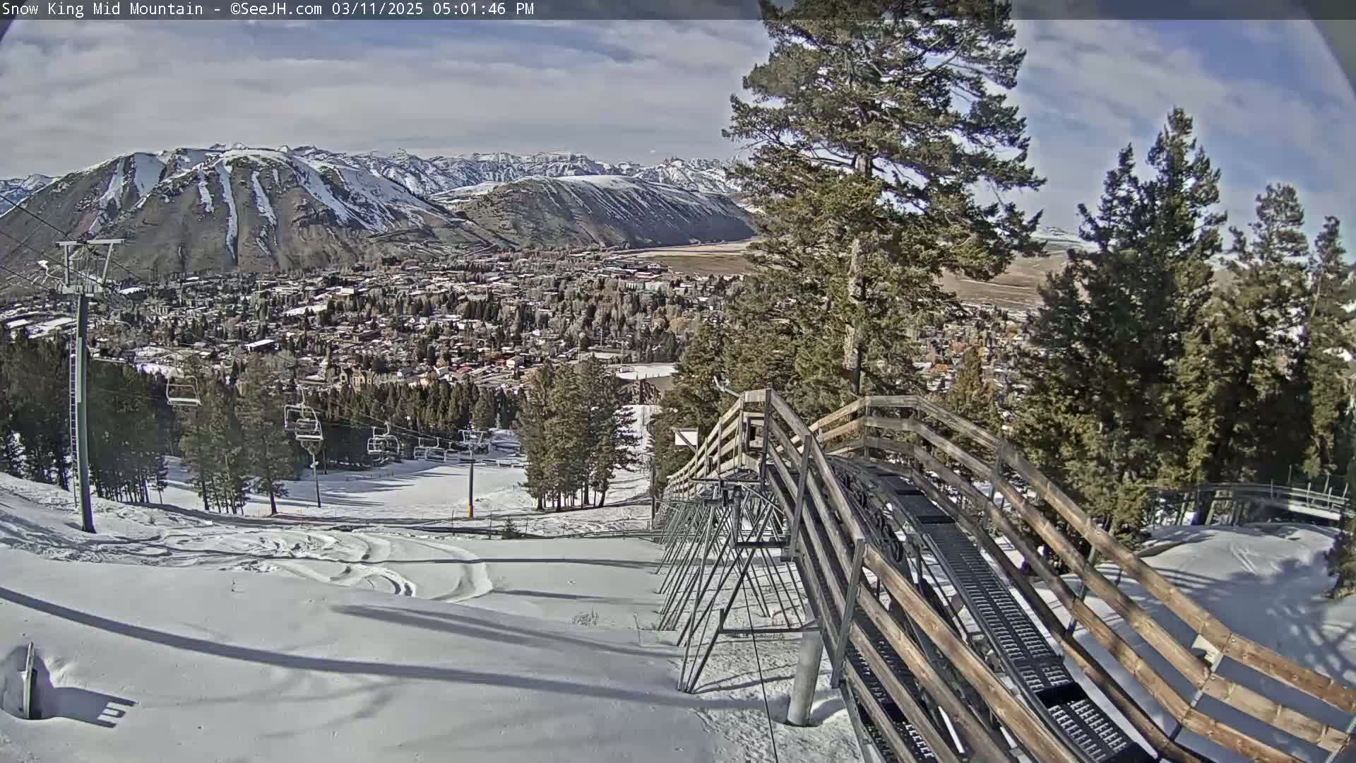 A snow-covered ski resort, with a mountain town visible in the distance, under partly sunny skies, is seen from a ski lift.