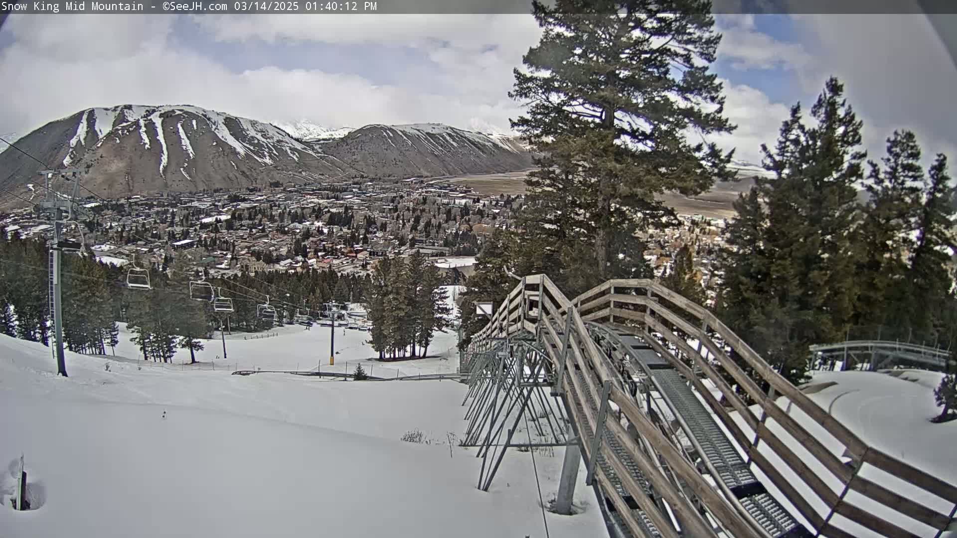 A mountain town is visible below a snowy mountainside, with a wooden alpine coaster descending towards it under partly cloudy skies.