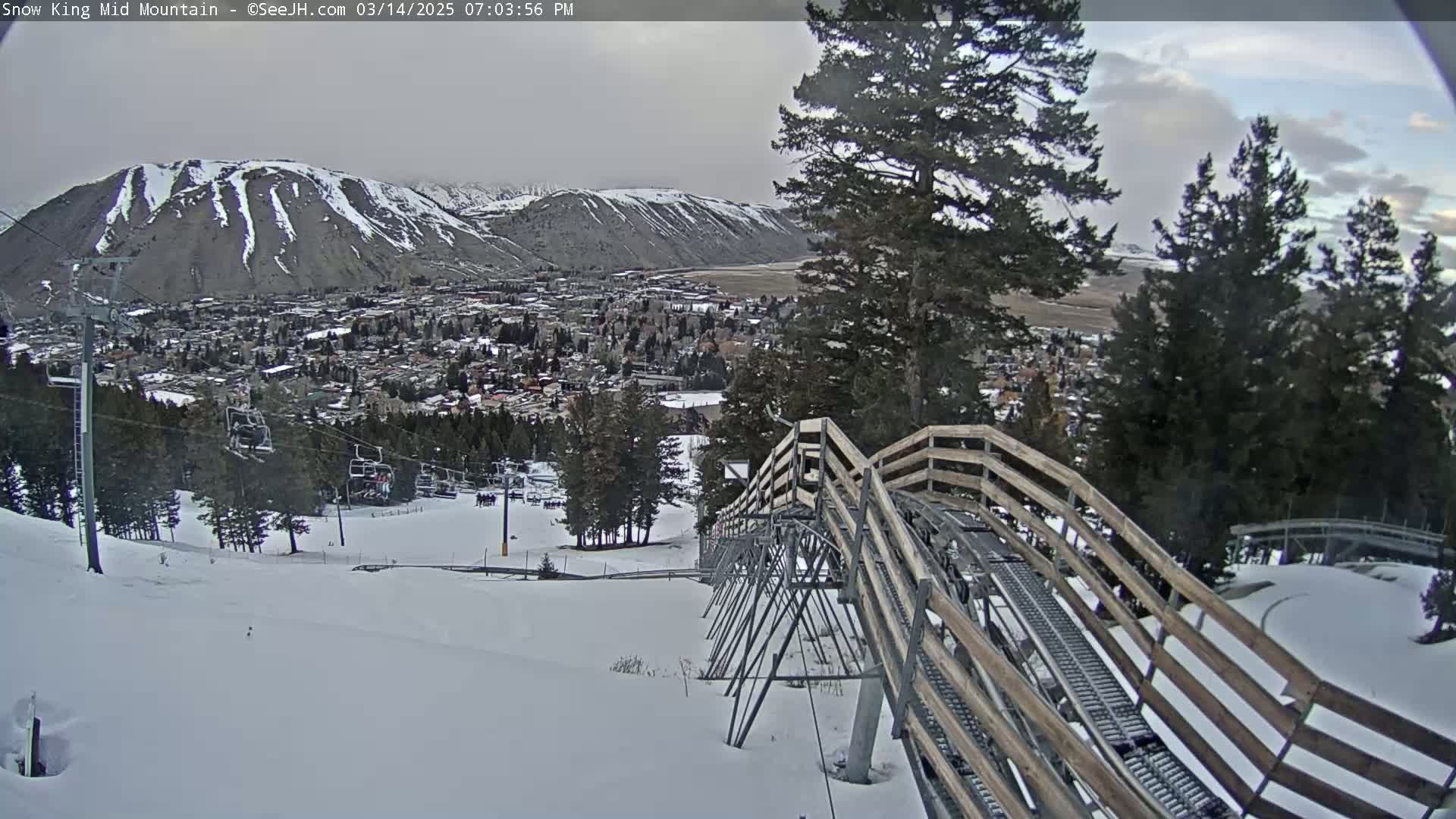 A snow-covered mountain slope with a wooden alpine coaster descends toward a town nestled in a valley under an overcast sky.