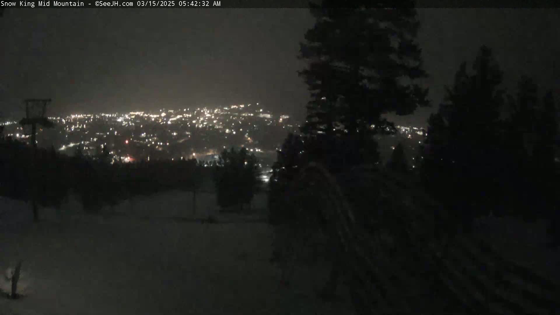 A nighttime view from a snowy, mountainous area shows a distant city illuminated with lights.