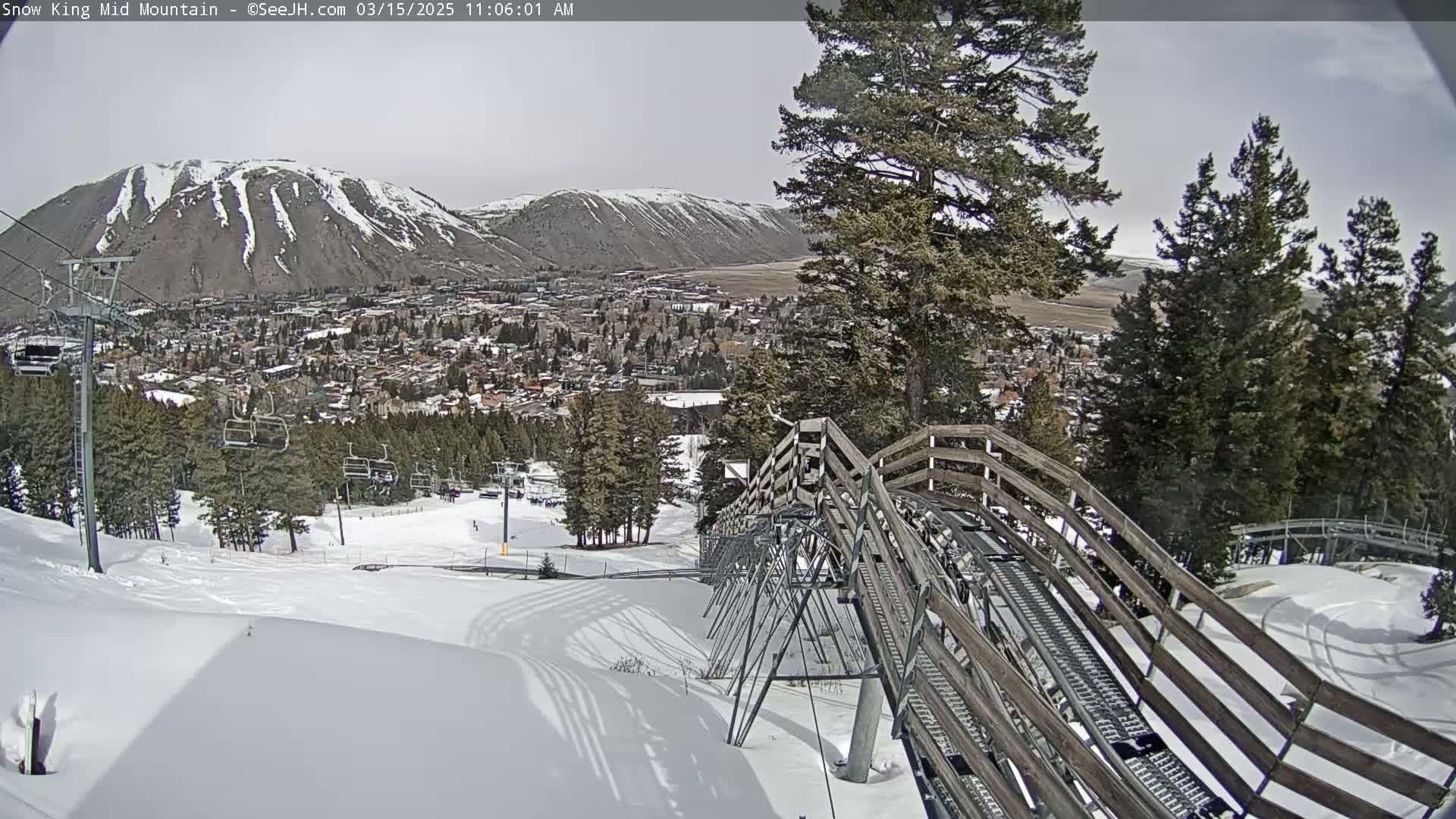 A snow-covered mountainside with a wooden alpine coaster leading down towards a town visible in the distance under partly cloudy skies.