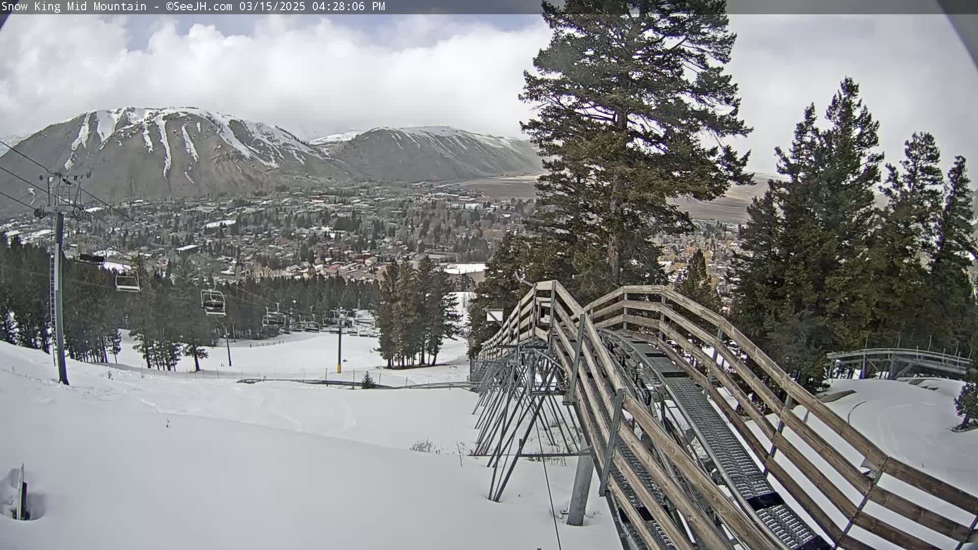 A snow-covered mountainside with a wooden alpine coaster descending towards a town nestled in a valley under a cloudy sky.