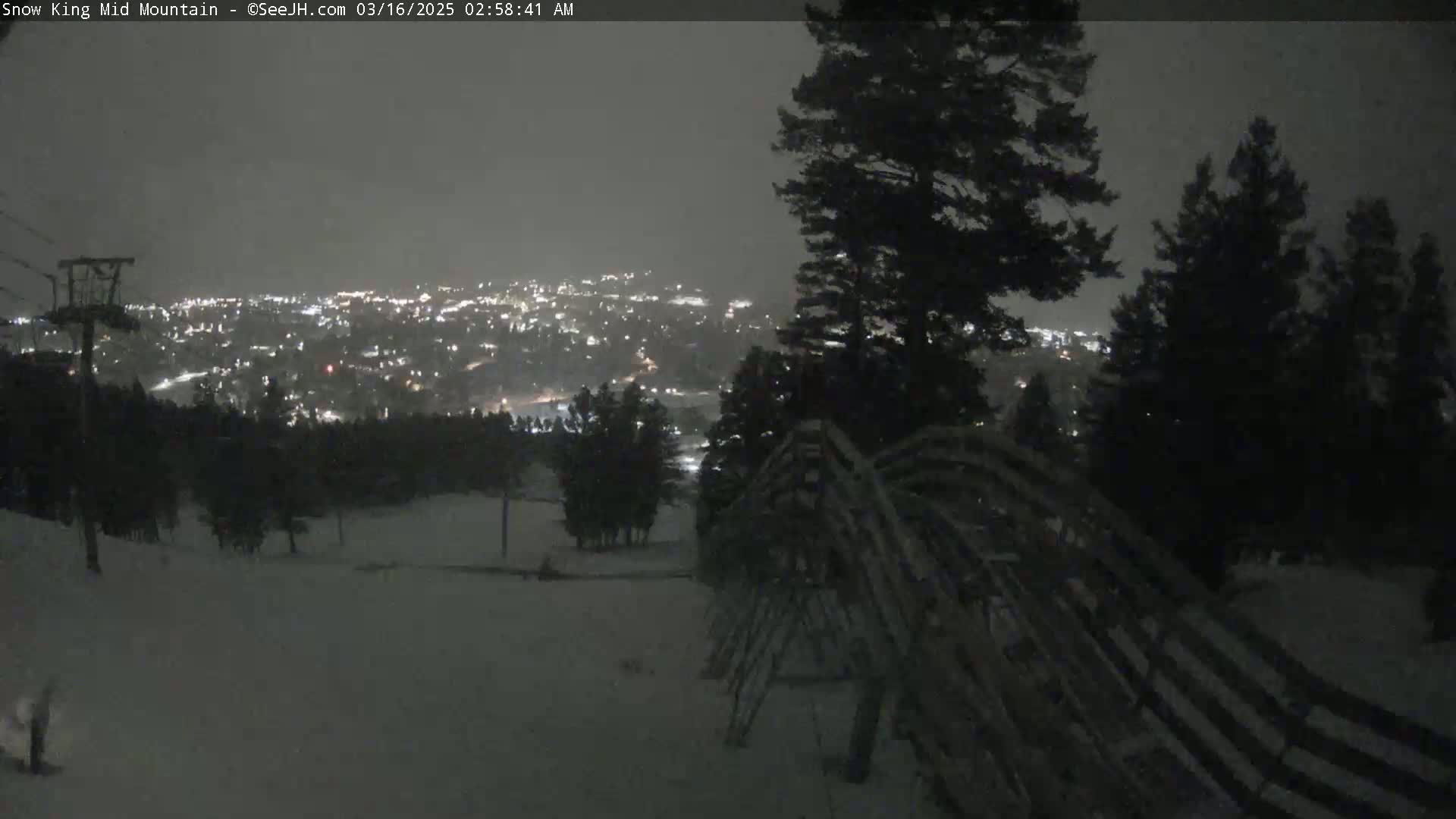 A nighttime, snowy scene shows a snow-covered slope overlooking a distant town illuminated by lights under a dark, overcast sky.