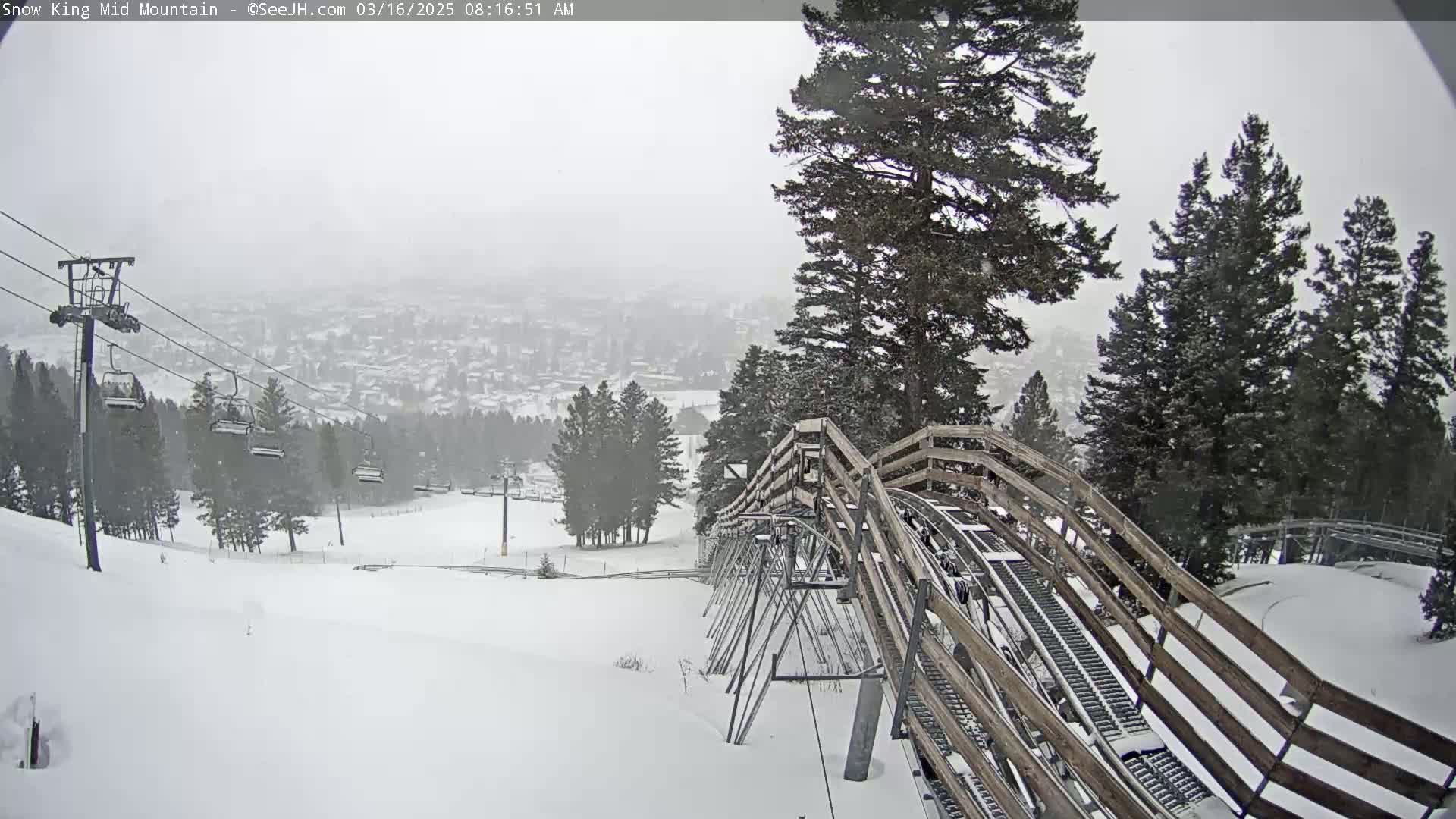 A snow-covered alpine coaster descends a hill towards a snowy town, with a ski lift and snow-covered evergreens in the background during a light snowstorm.