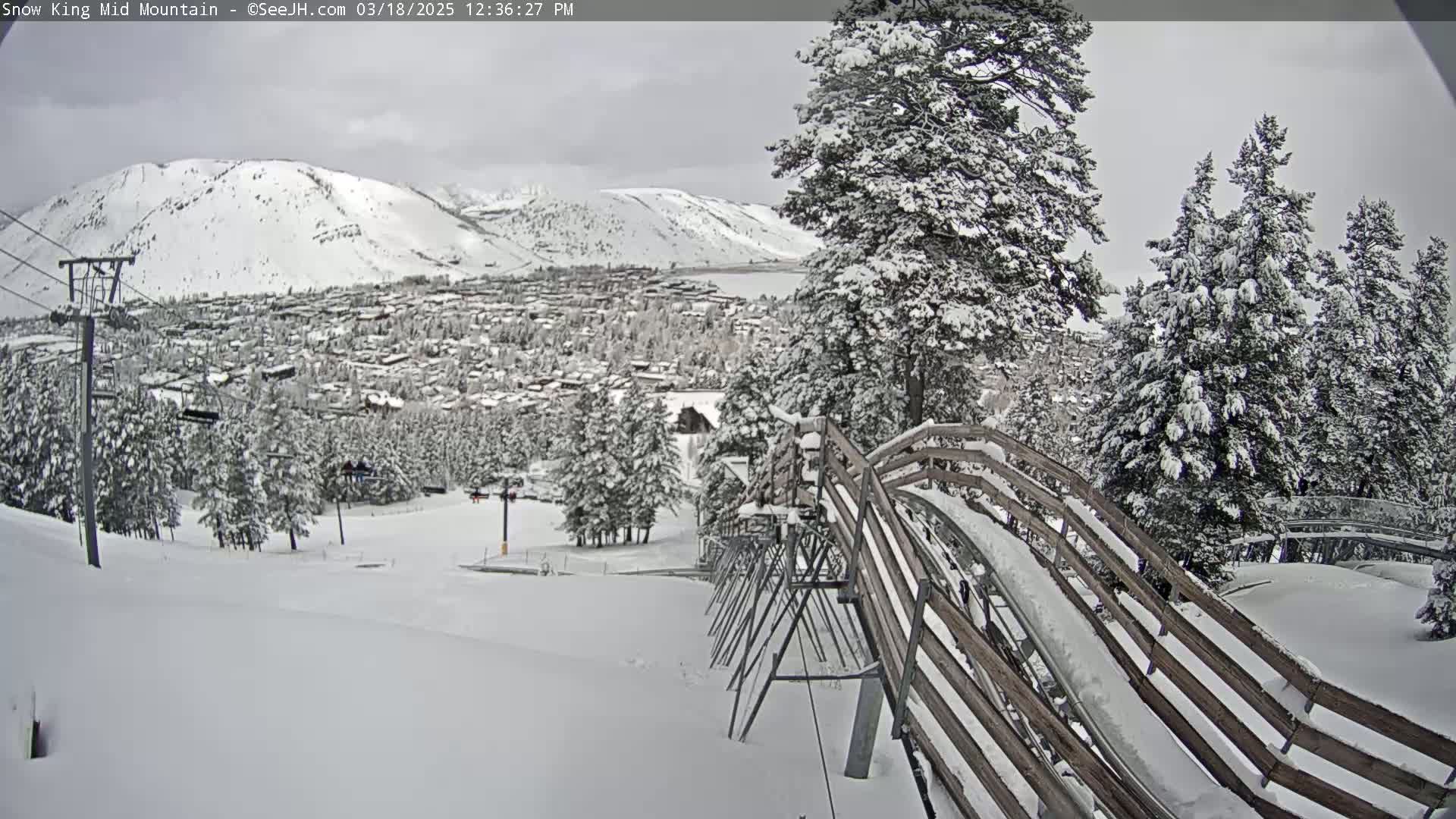 A snow-covered mountain town is visible in the distance, viewed from a snow-covered alpine coaster on a cloudy day.