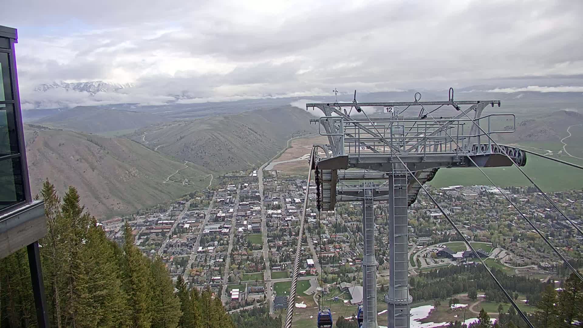 A mountain town is visible below a chairlift on an overcast day, with snow-capped mountains in the distance.