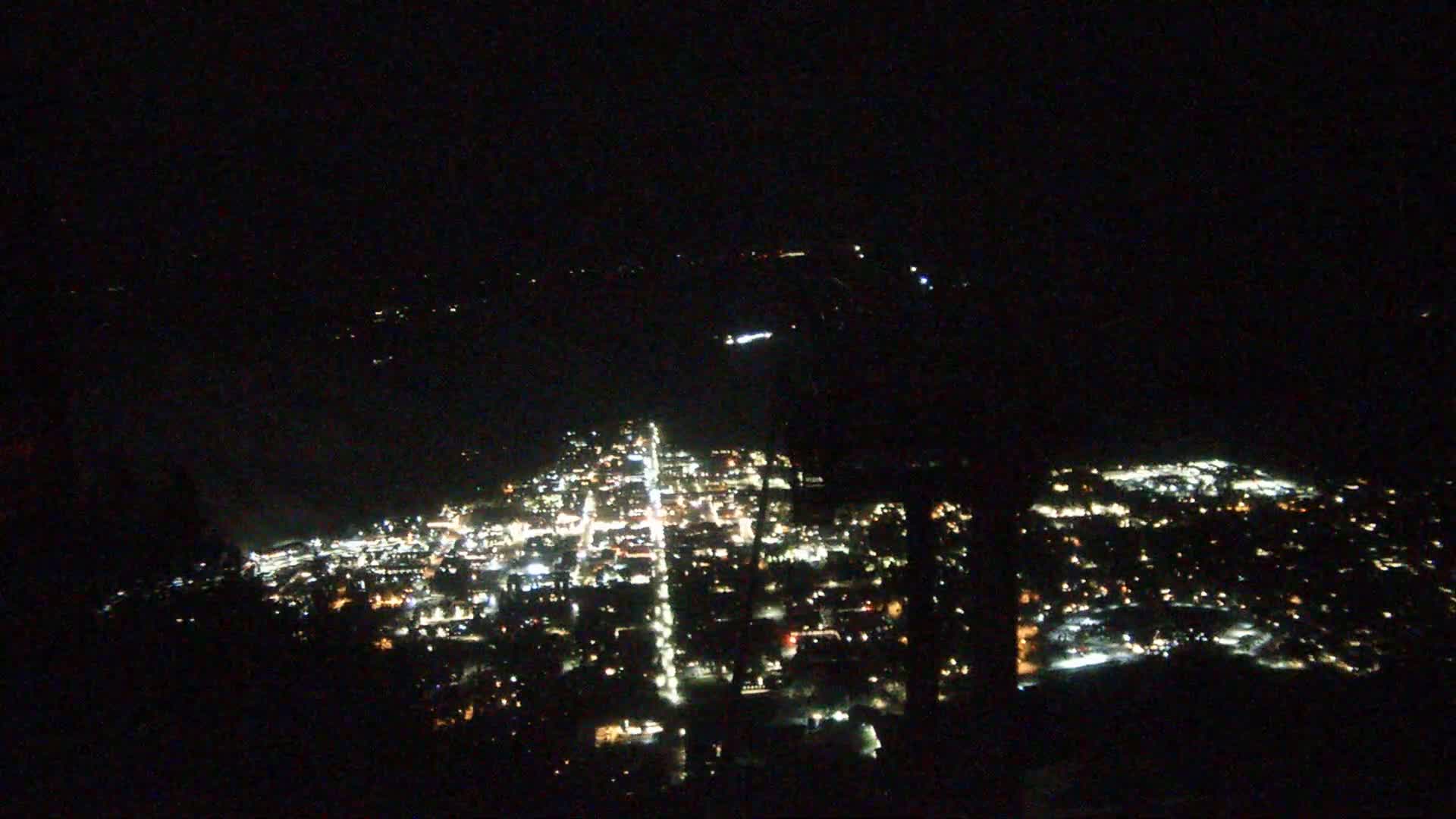 A nighttime, high-angle view of a city illuminated by streetlights and building lights, extending to a dark body of water in the distance.