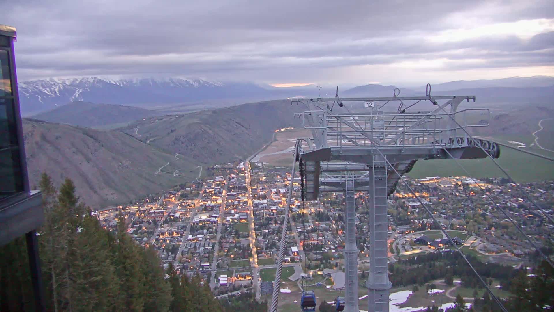 A nighttime view from a chairlift overlooking a valley town illuminated by lights, under a cloudy sky with snow-capped mountains in the distance.