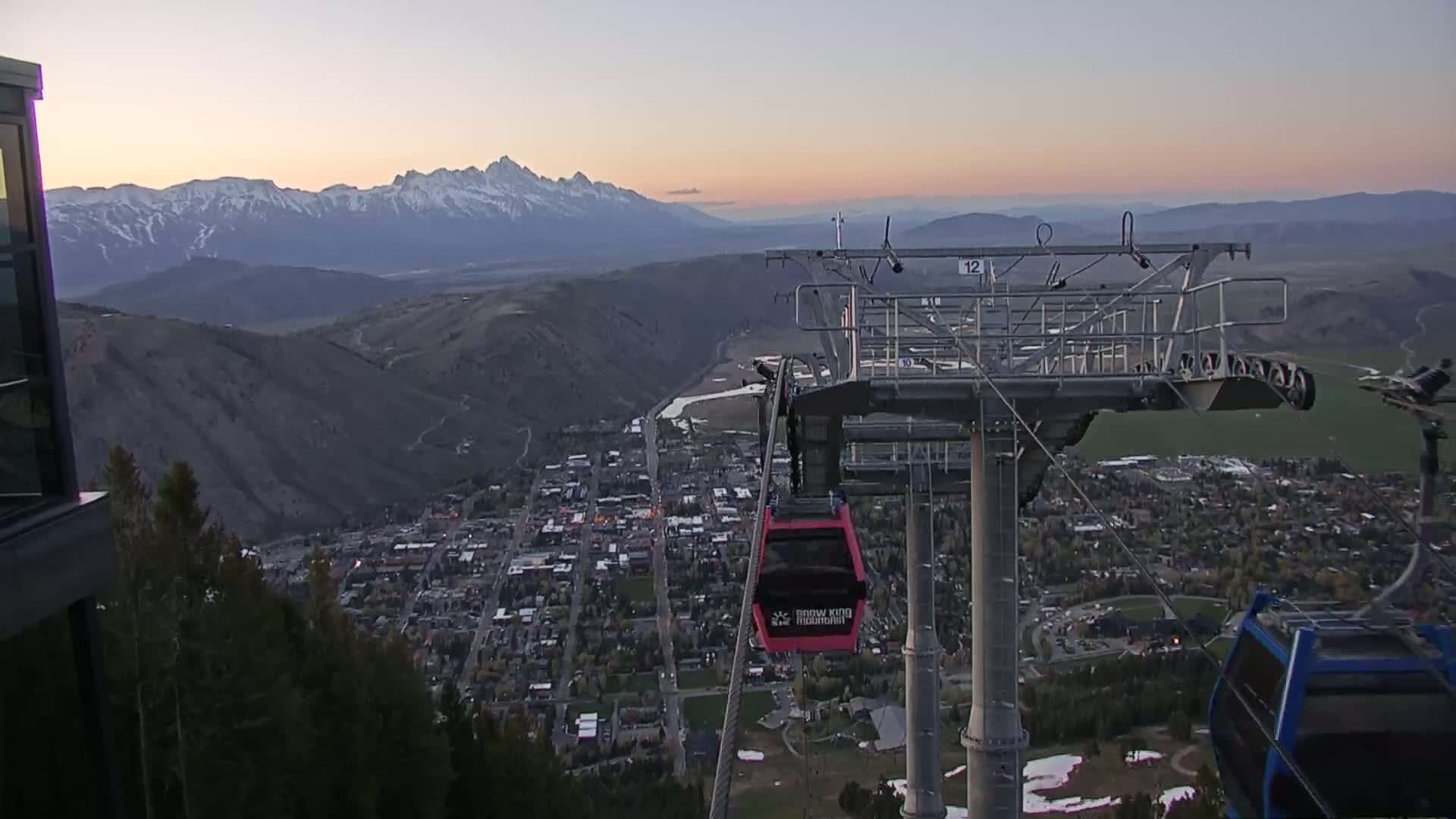 A chairlift ascends above a town nestled in a valley, overlooked by snow-capped mountains under a twilight sky.