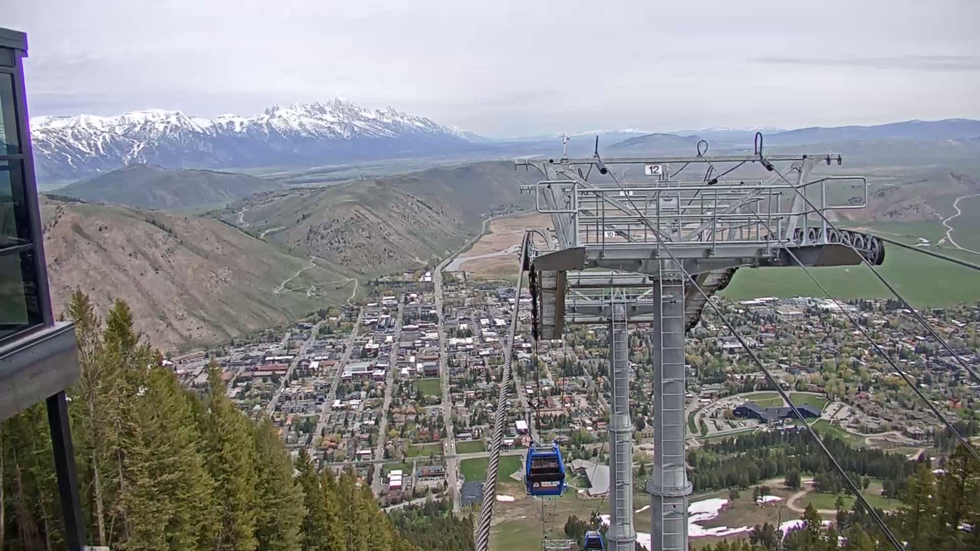 A chairlift ascends a mountain overlooking a valley town nestled beneath snow-capped mountains on a partly cloudy day.