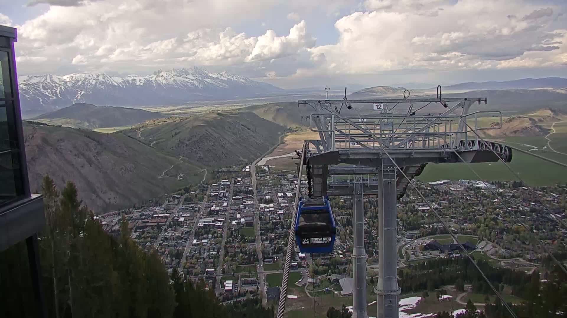 A mountain town is seen from a gondola on a partly cloudy day, with snow-capped mountains in the background.
