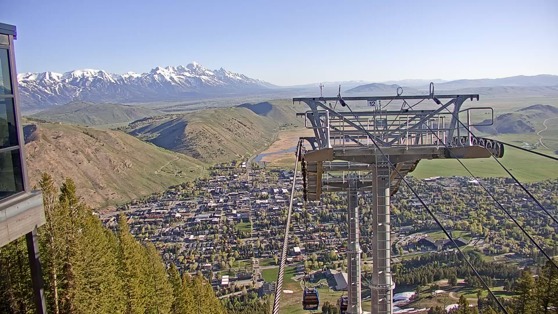 A chairlift overlooks a town nestled in a valley, with snow-capped mountains in the background on a clear, sunny day.