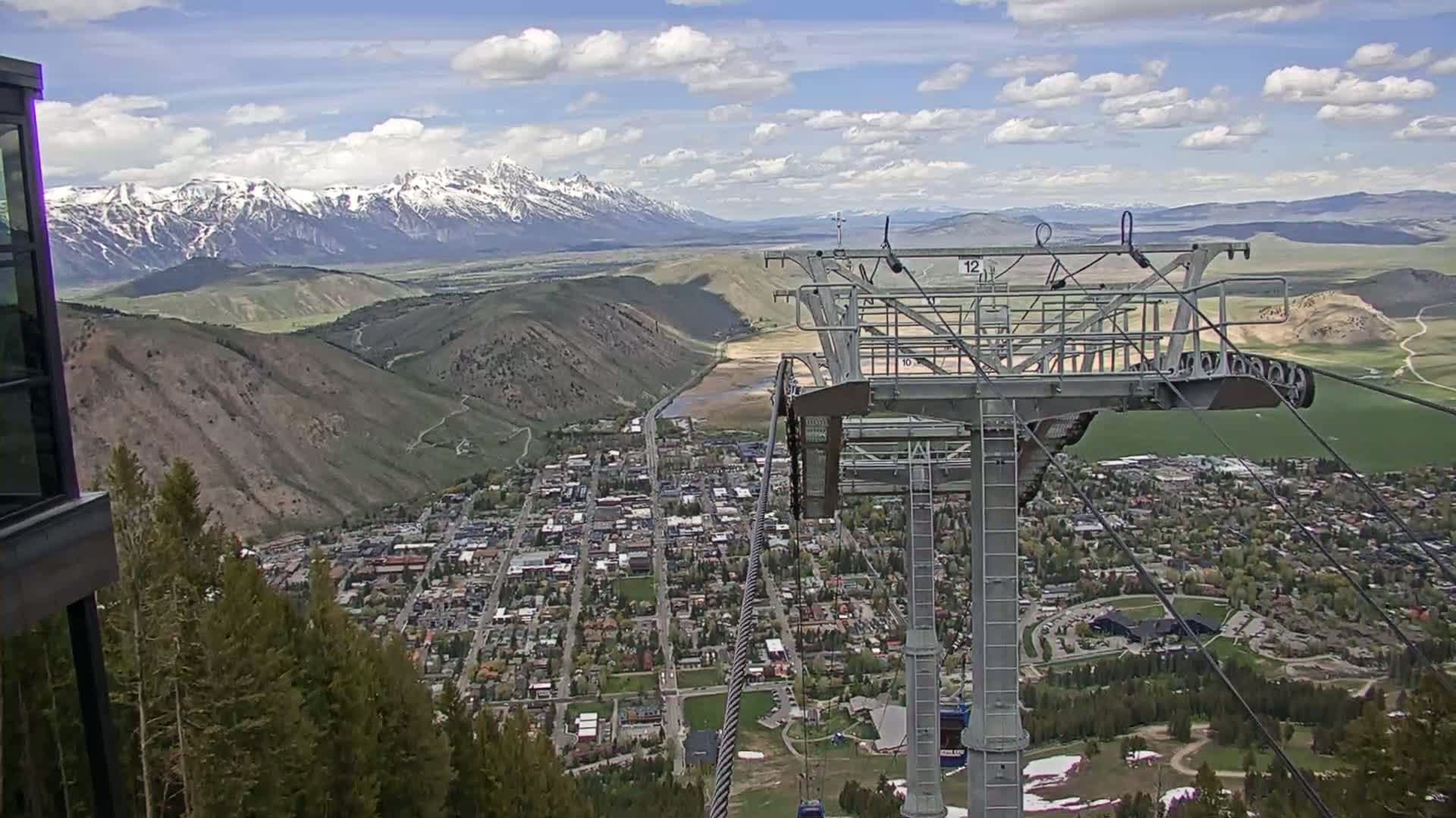 A partly cloudy day shows a mountain town nestled in a valley below a snow-capped mountain range, viewed from a ski lift.