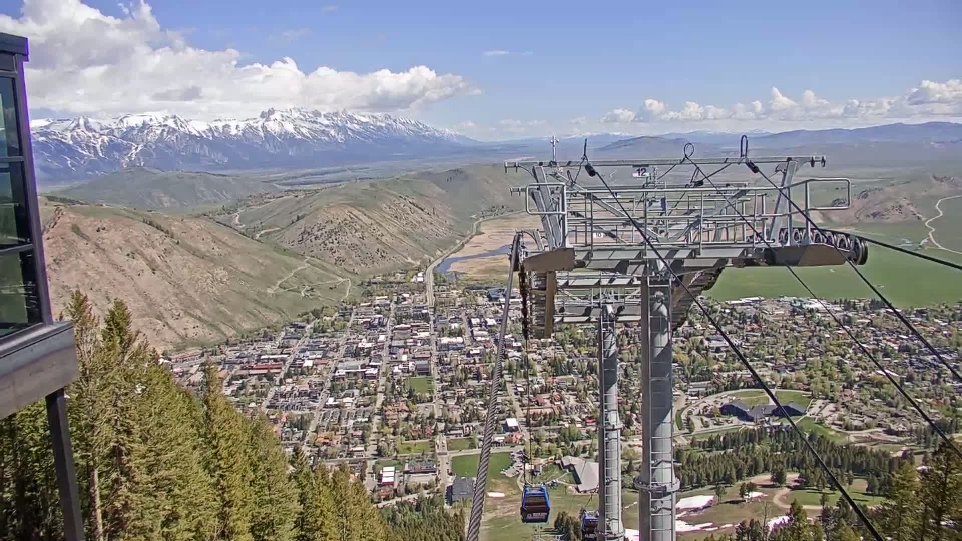 A mountain town nestled in a valley, viewed from a ski lift on a partly cloudy day with snow-capped mountains in the background.