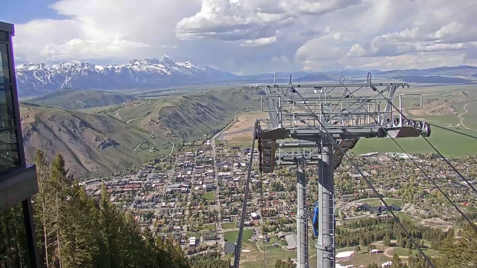A chairlift ascends above a mountain town nestled in a valley, with snow-capped mountains in the background under a partly cloudy sky.