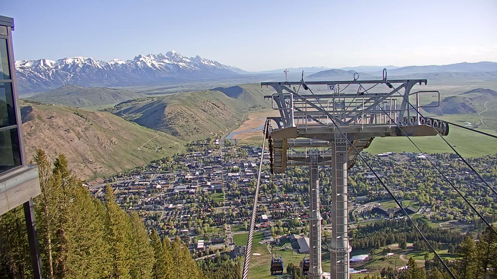 A chairlift carrying cable ascends a mountain overlooking a town nestled in a valley, with snow-capped mountains in the background under a clear sky.