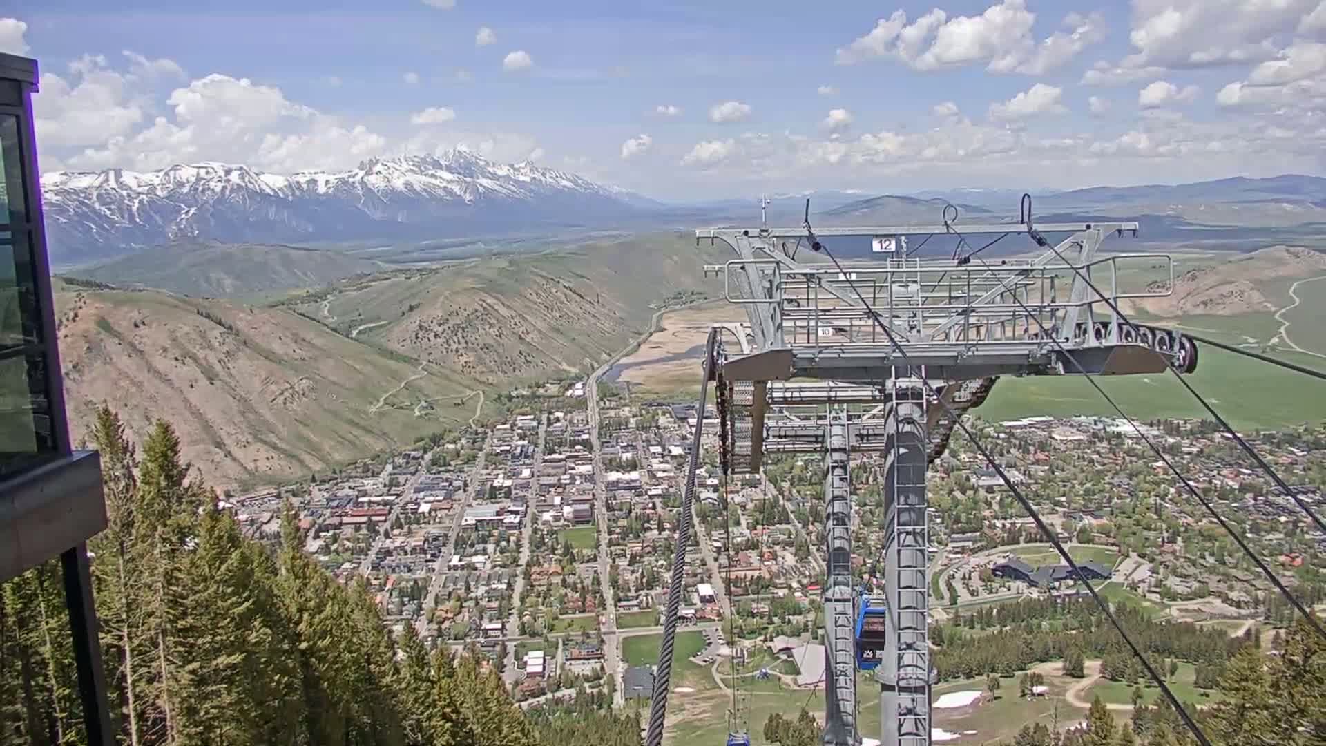 A chairlift ascends a mountain overlooking a town nestled in a valley, with snow-capped mountains in the background under a partly cloudy sky.