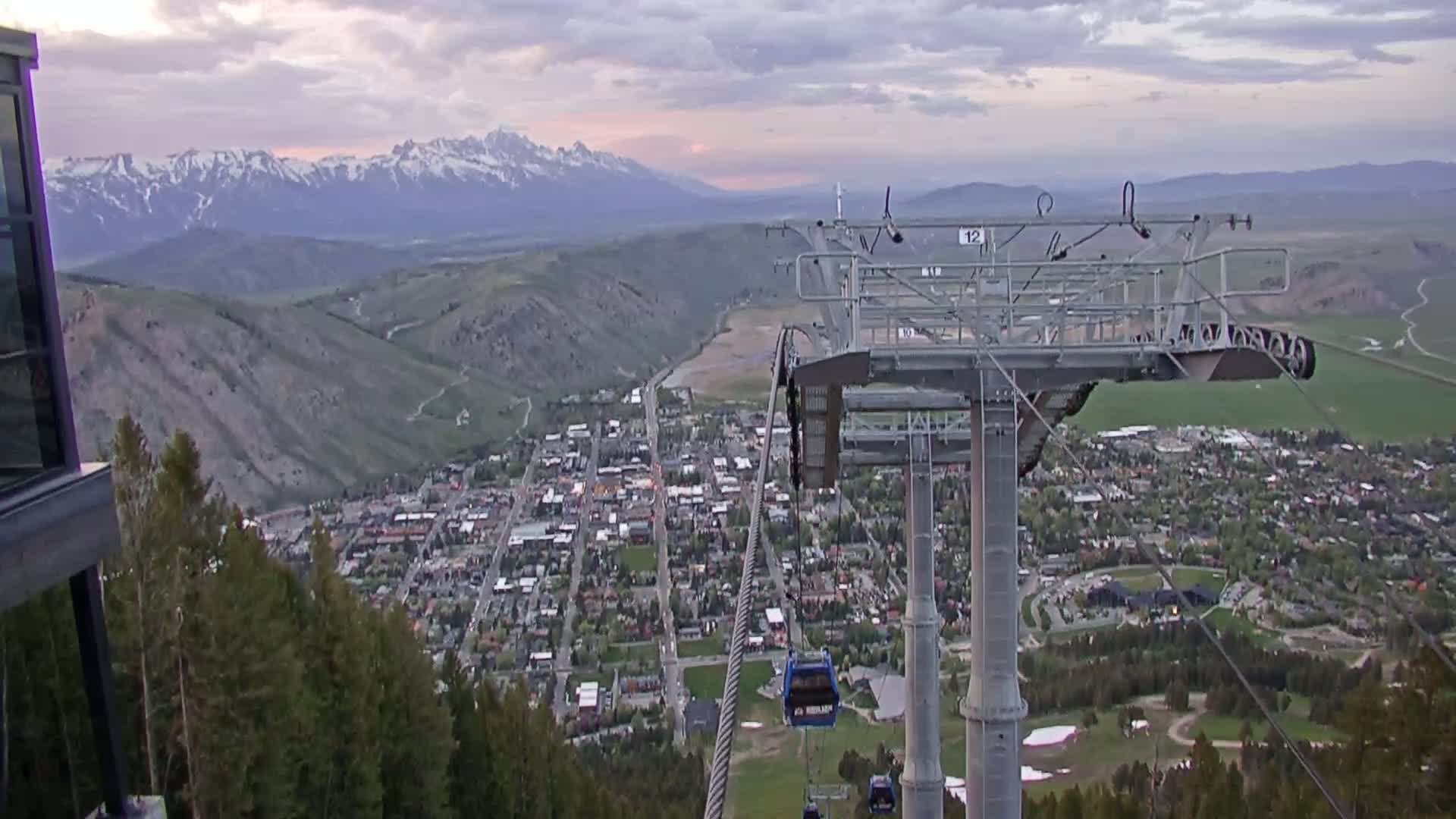 A chairlift ascends a mountain overlooking a valley town and snow-capped mountains in the distance under a cloudy, dusky sky.
