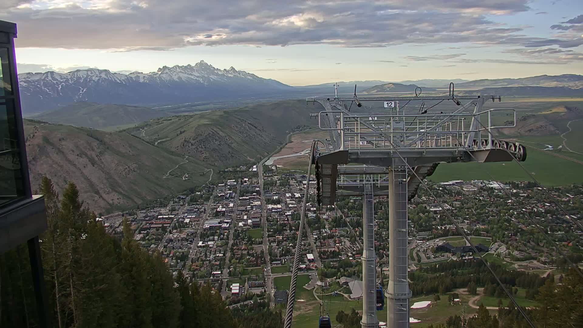 A chairlift ascends a mountain overlooking a town nestled in a valley, with snow-capped mountains in the background under a partly cloudy sky.