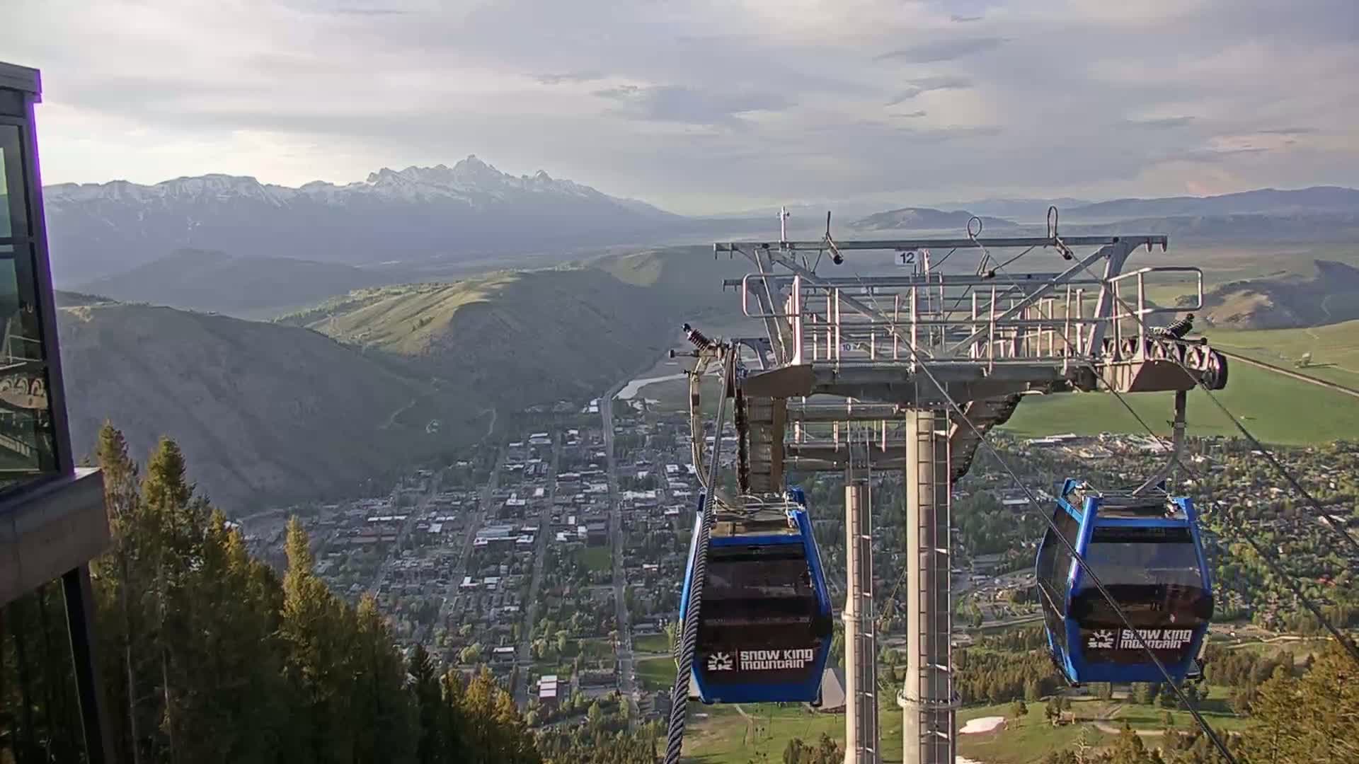 A mountain town is visible below a chairlift ascending a mountain on a partly cloudy day, with snow-capped mountains in the background.