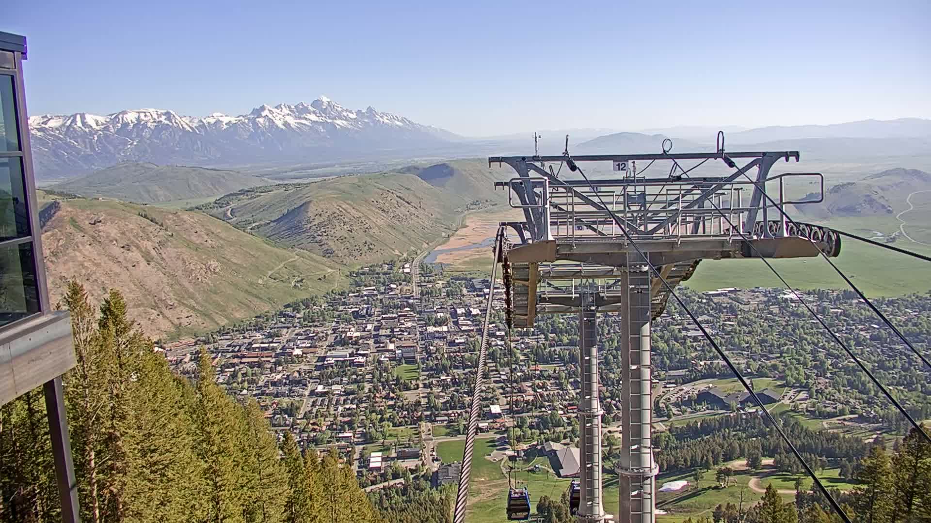A mountain town lies below a chairlift on a clear, sunny day, with snow-capped mountains visible in the distance.