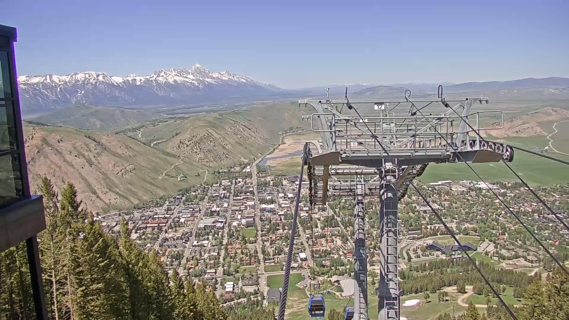A chairlift ascends a mountainside overlooking a valley town nestled beneath snow-capped mountains on a clear, sunny day.