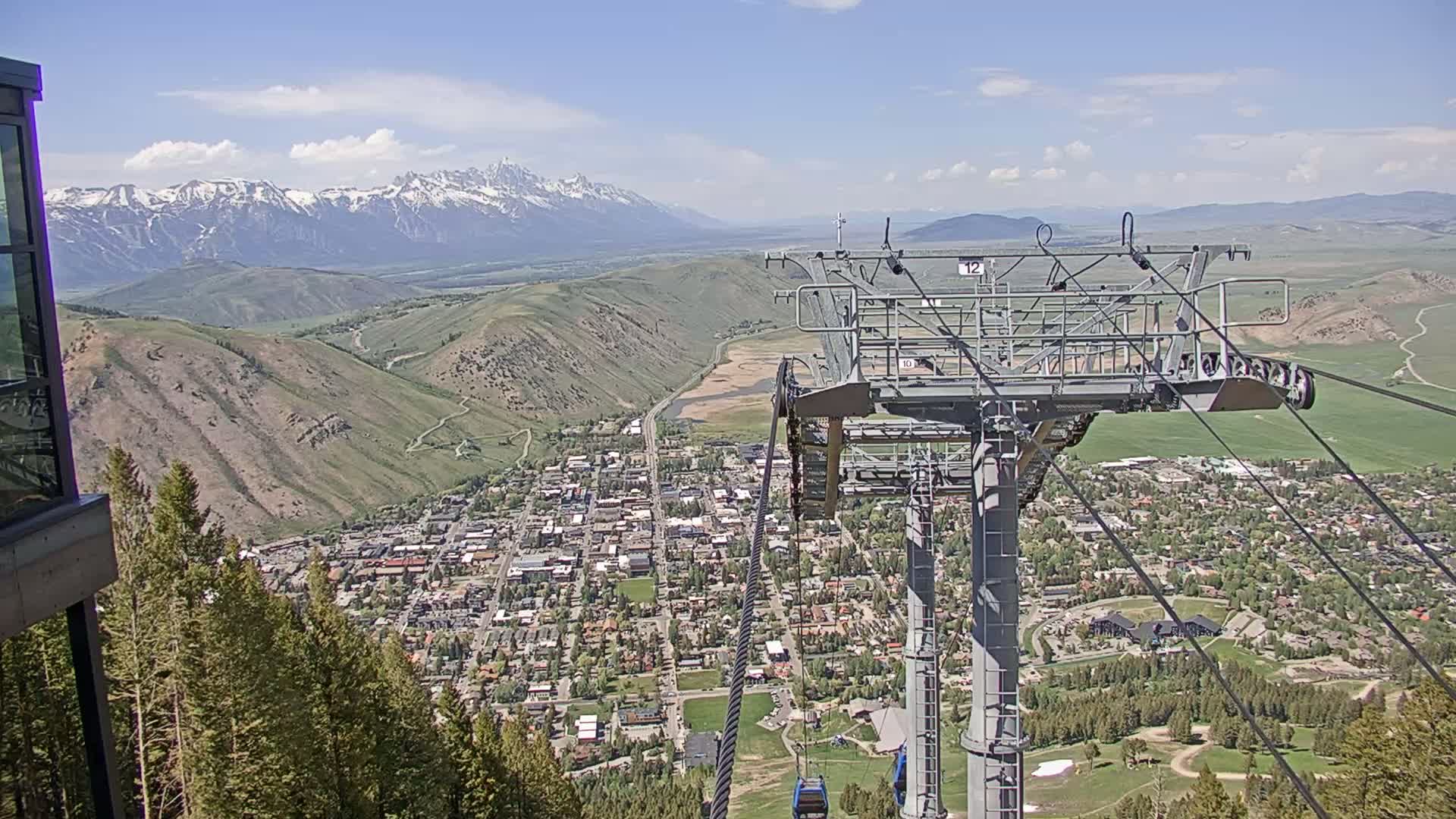 A chairlift overlooks a town nestled in a valley, with snow-capped mountains in the background under a mostly sunny sky.