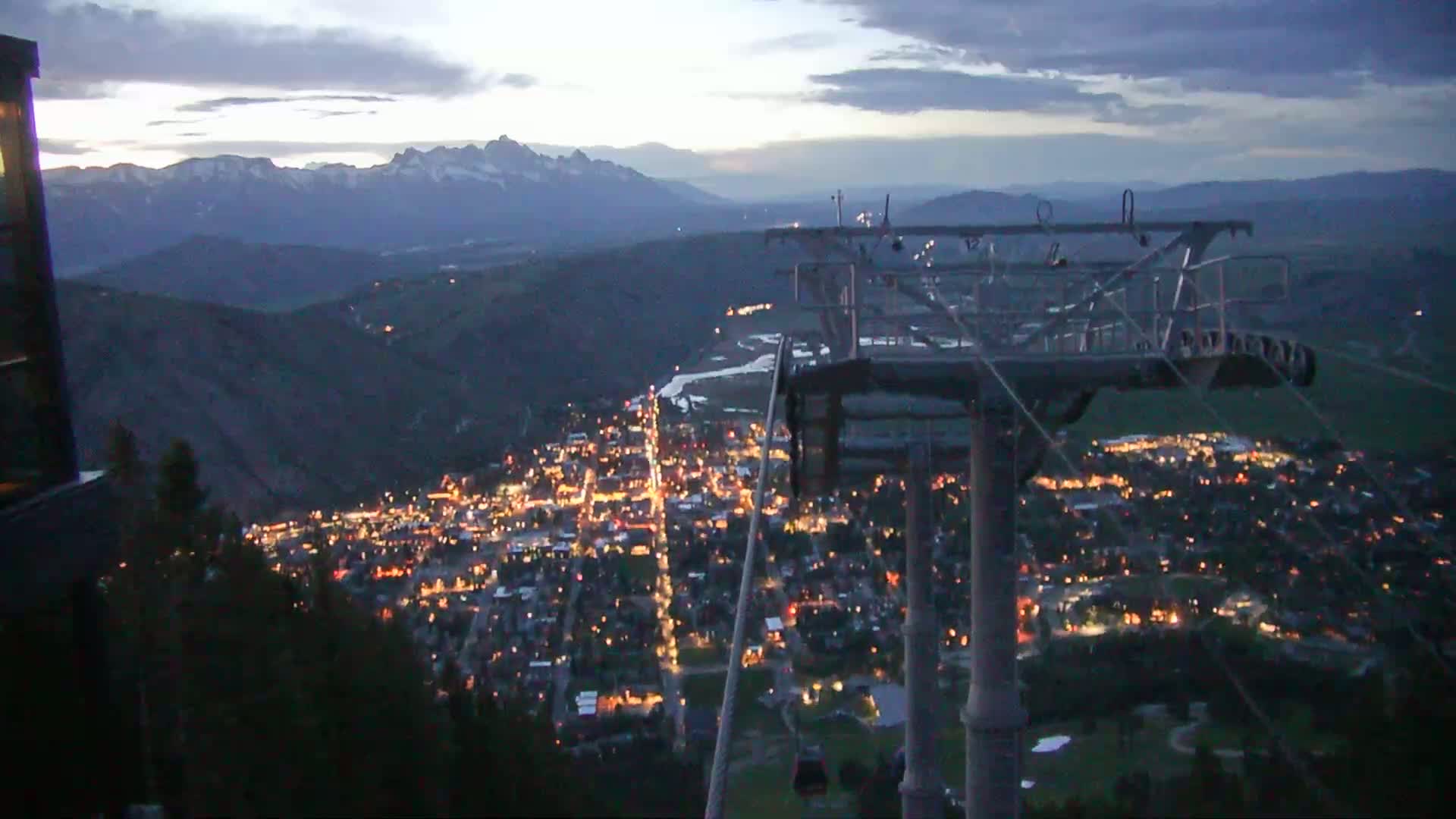 A nighttime view from a chairlift shows a valley town illuminated with lights, nestled between mountains under a partly cloudy sky.