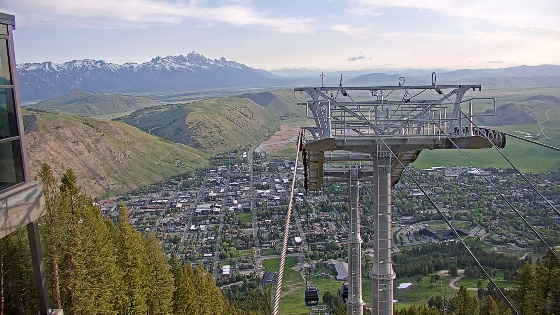 A chairlift carrying cable ascends a mountain overlooking a valley town nestled beneath snow-capped mountains on a partly cloudy day.