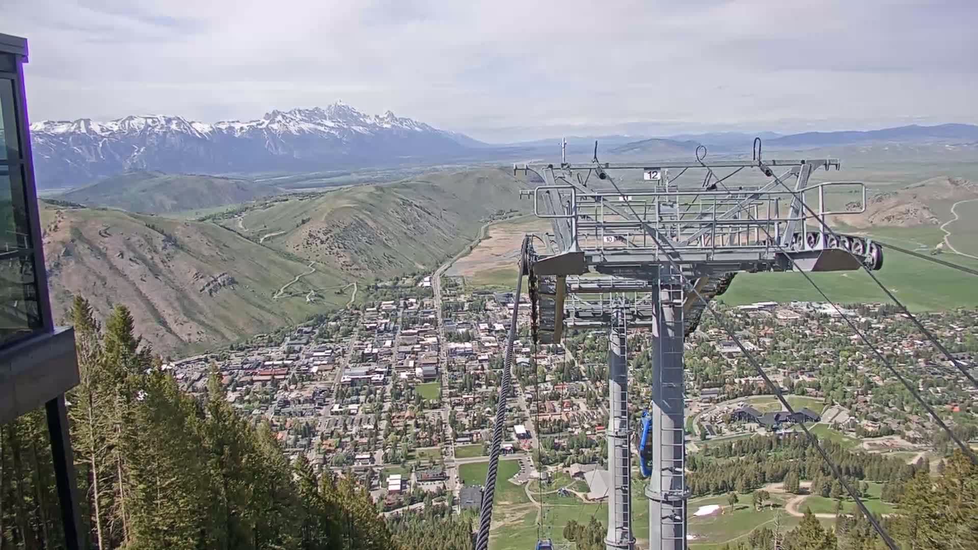 A chairlift overlooks a town nestled in a valley surrounded by green hills and snow-capped mountains under a partly cloudy sky.