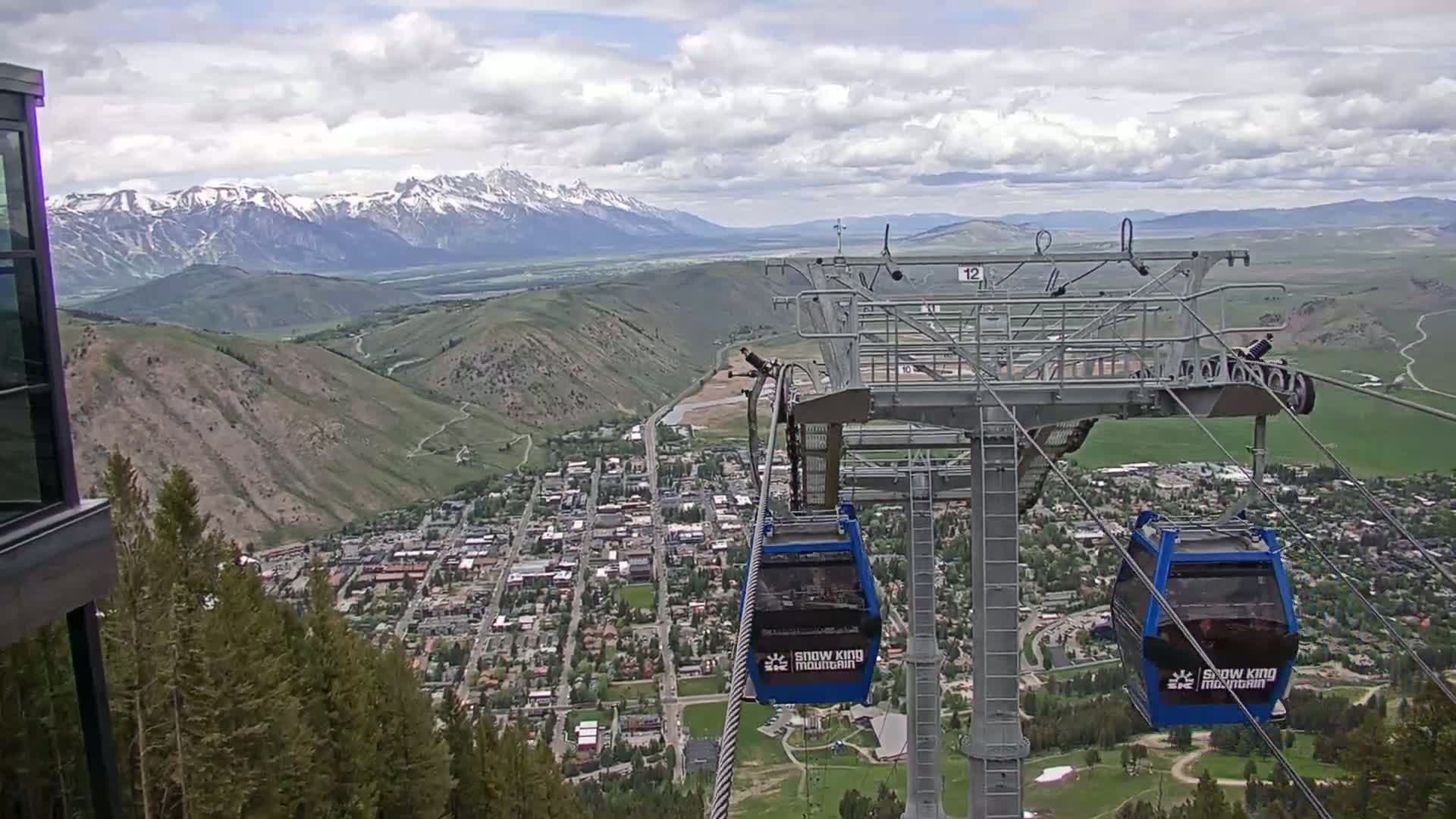 A partly cloudy day shows a mountain town nestled in a valley below a chairlift, with snow-capped mountains in the background.