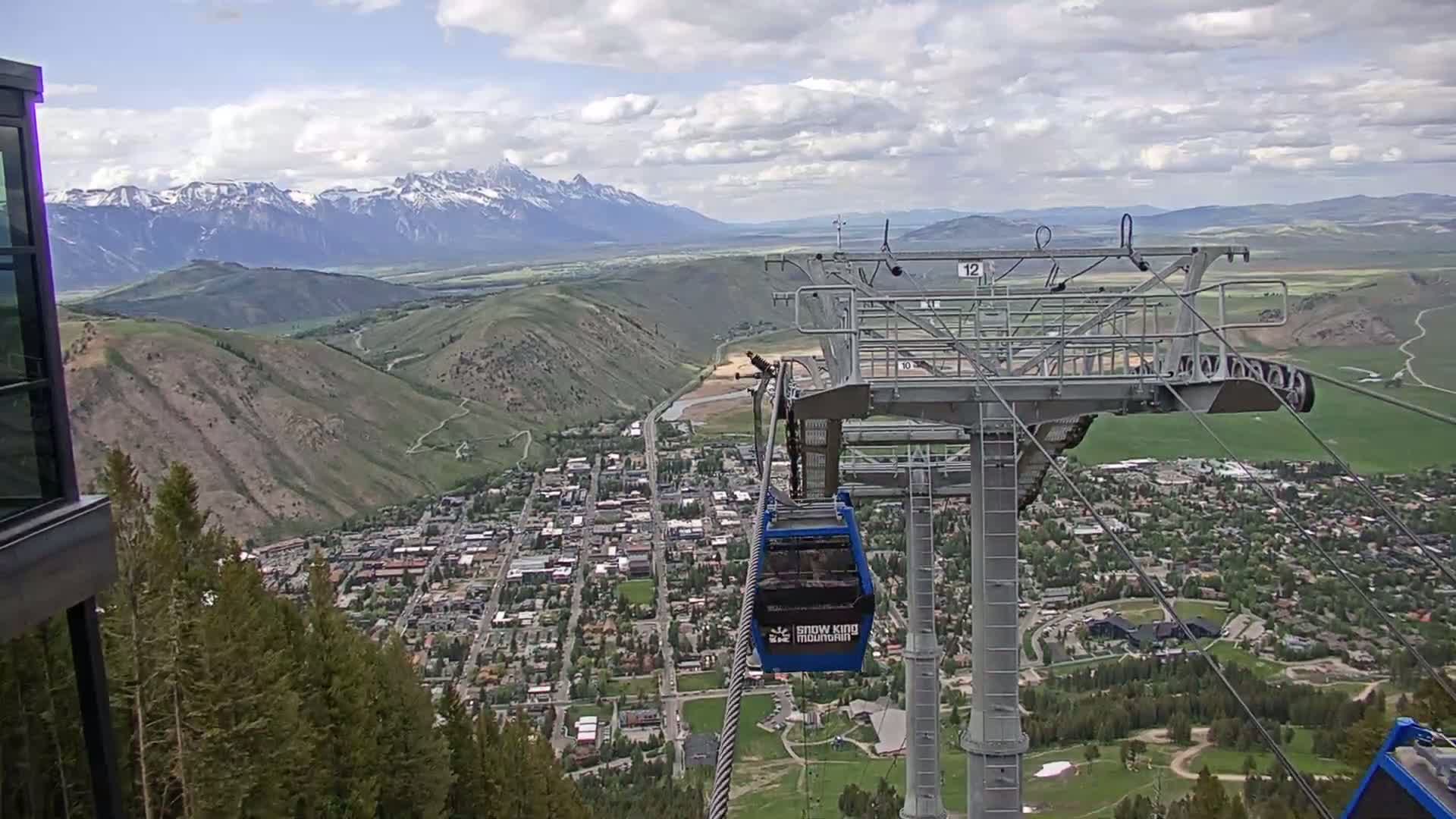 A partially cloudy day shows a mountain town nestled in a valley below a ski lift, with snow-capped mountains visible in the distance.