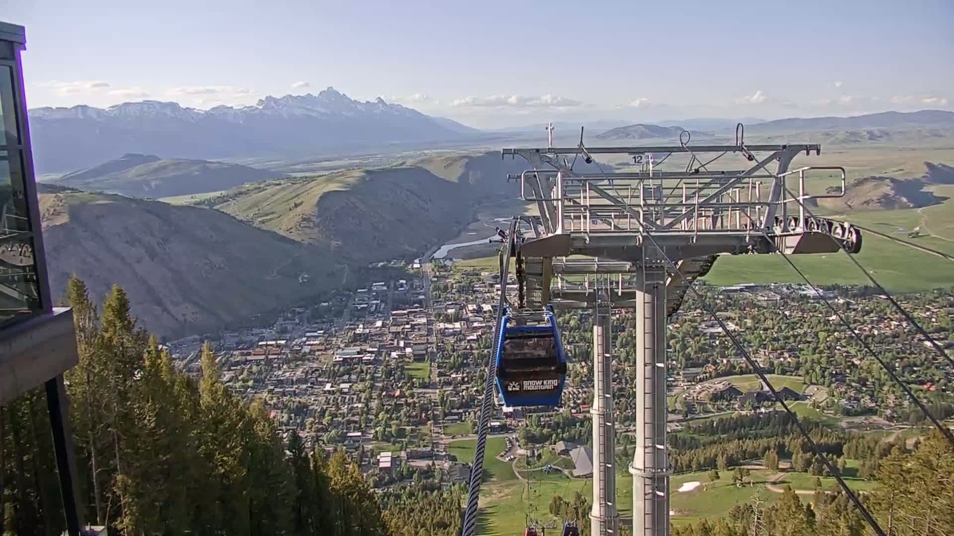 A chairlift gondola ascends above a town nestled in a valley, with snow-capped mountains visible in the background under a mostly clear sky.