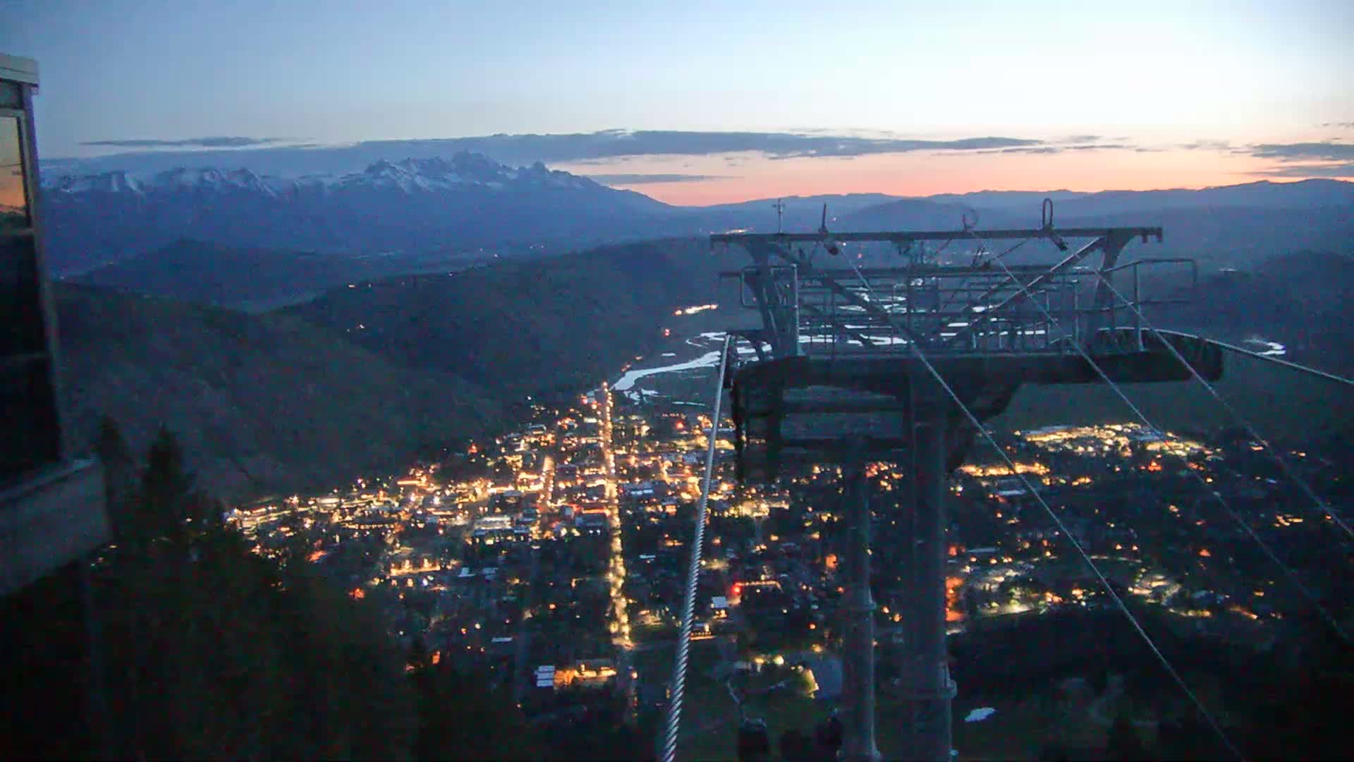 A nighttime view from a chairlift shows a valley town illuminated below, with snow-capped mountains in the distance under a twilight sky.