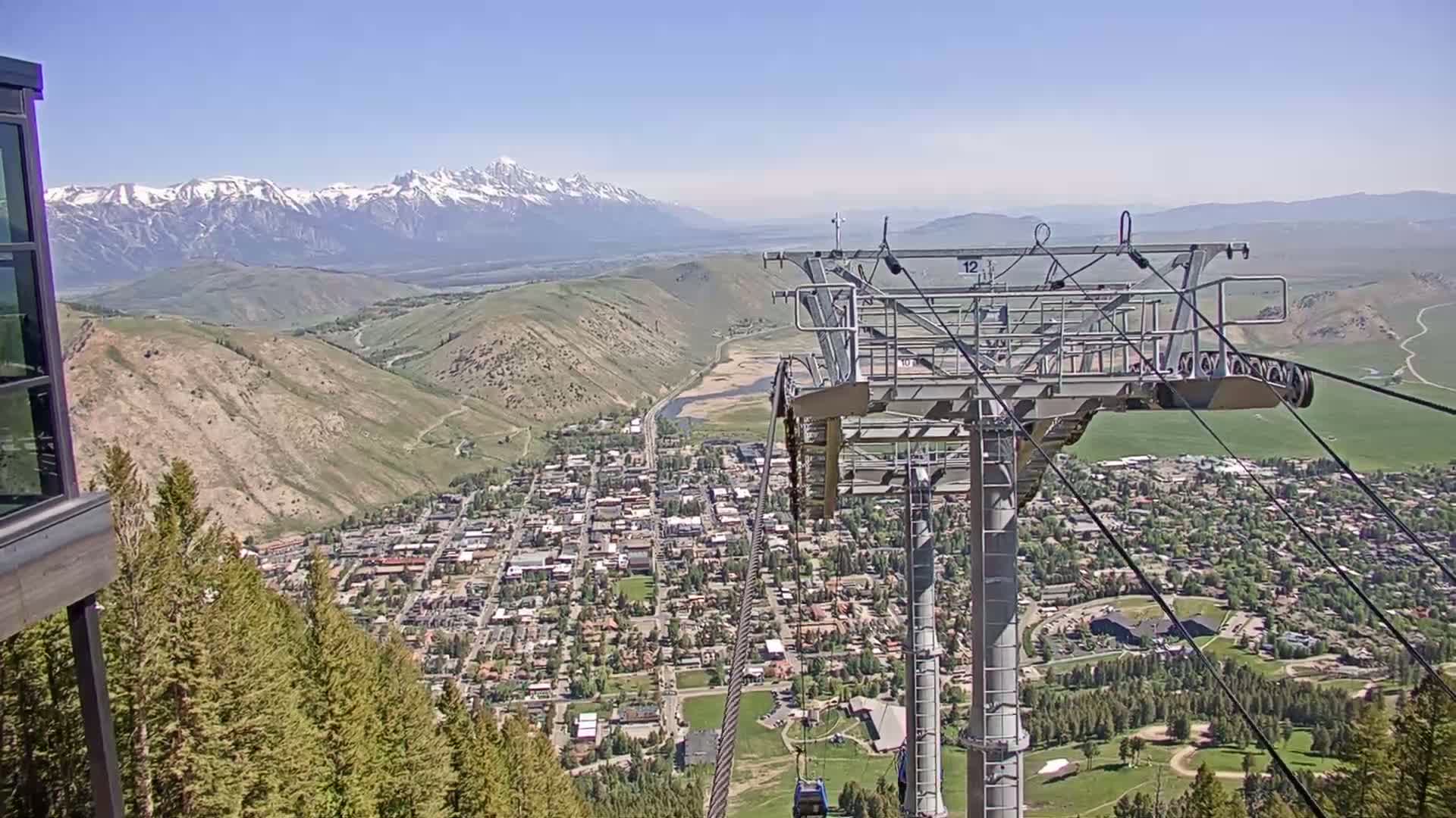 A mountain town is seen from a high vantage point on a clear, sunny day, with snow-capped mountains in the background and a chairlift in the foreground.