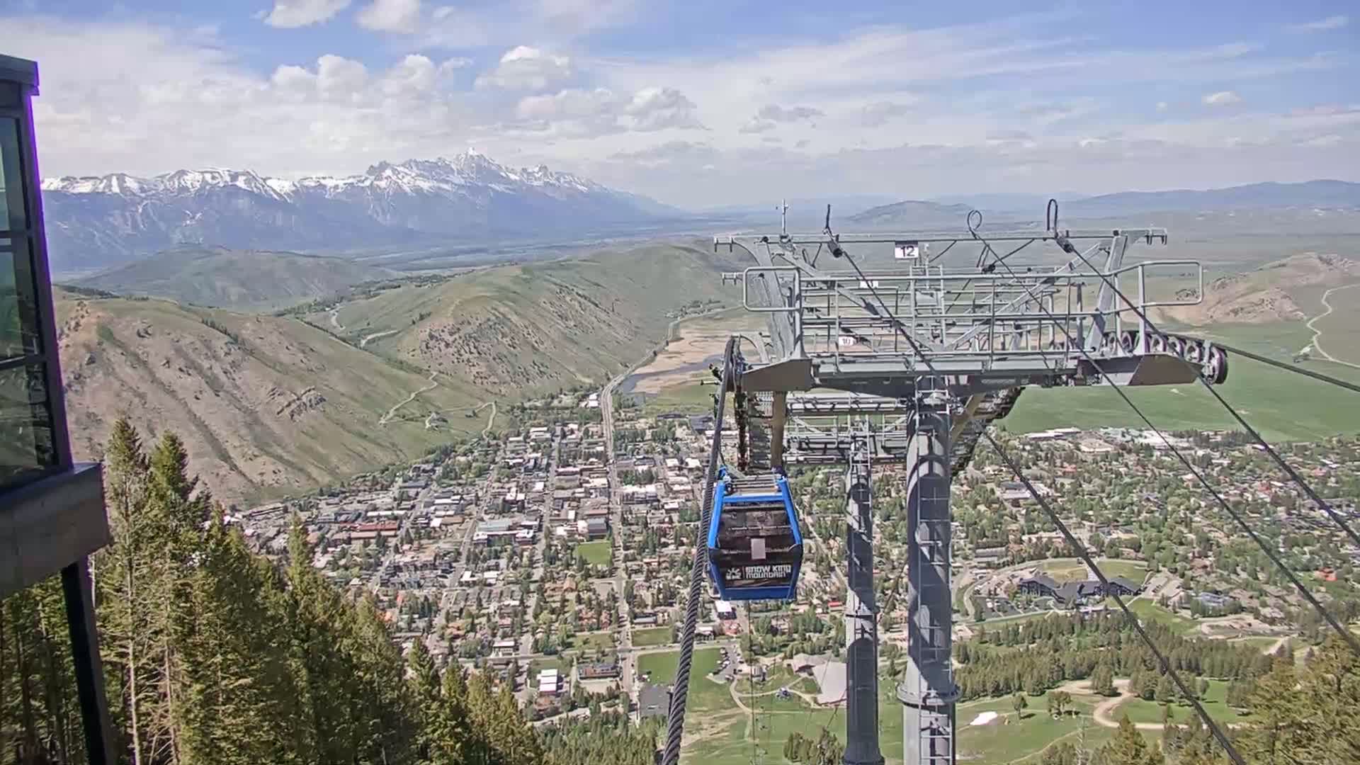 A chairlift gondola ascends above a mountain town nestled in a valley beneath snow-capped mountains on a partly cloudy day.