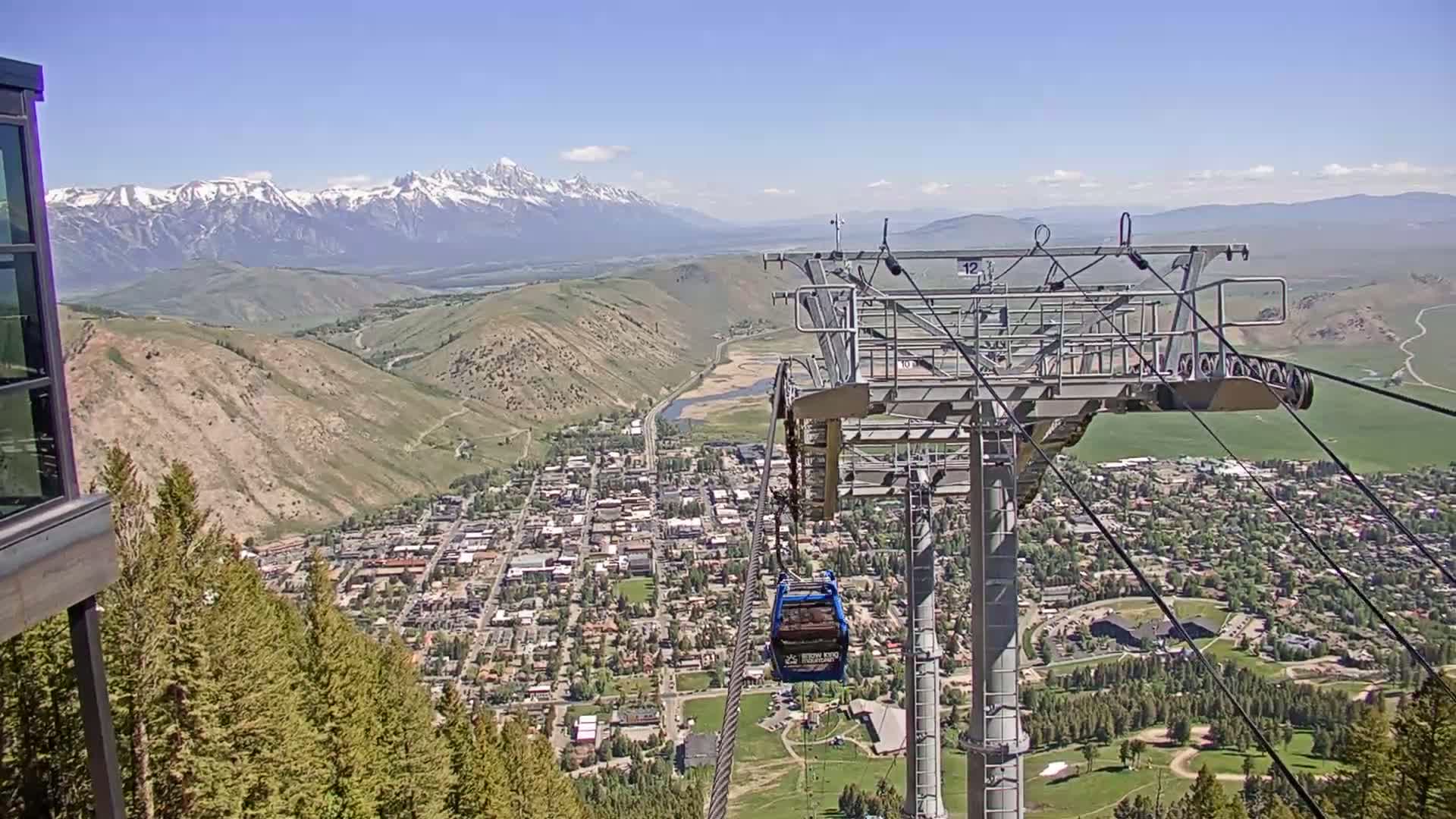 A sunny view from a chairlift shows a town nestled in a valley below snow-capped mountains.