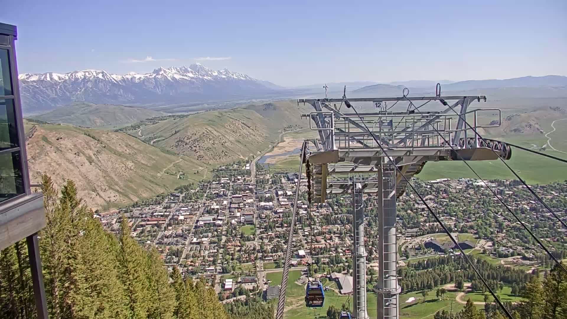 A mountain town sprawls below a chairlift on a sunny day, with snow-capped mountains in the background.