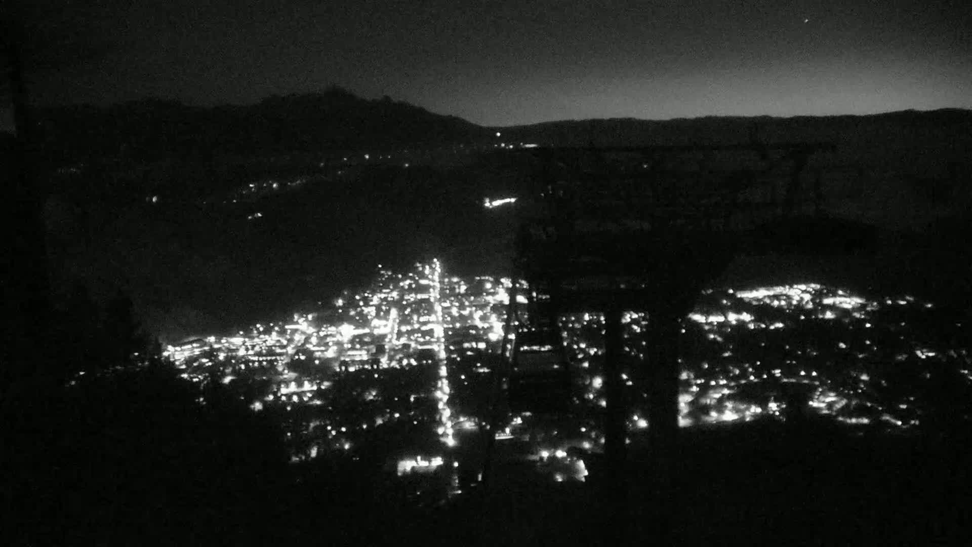 A nighttime view from an elevated vantage point shows a city illuminated below, nestled between dark mountains under a clear, dark sky.