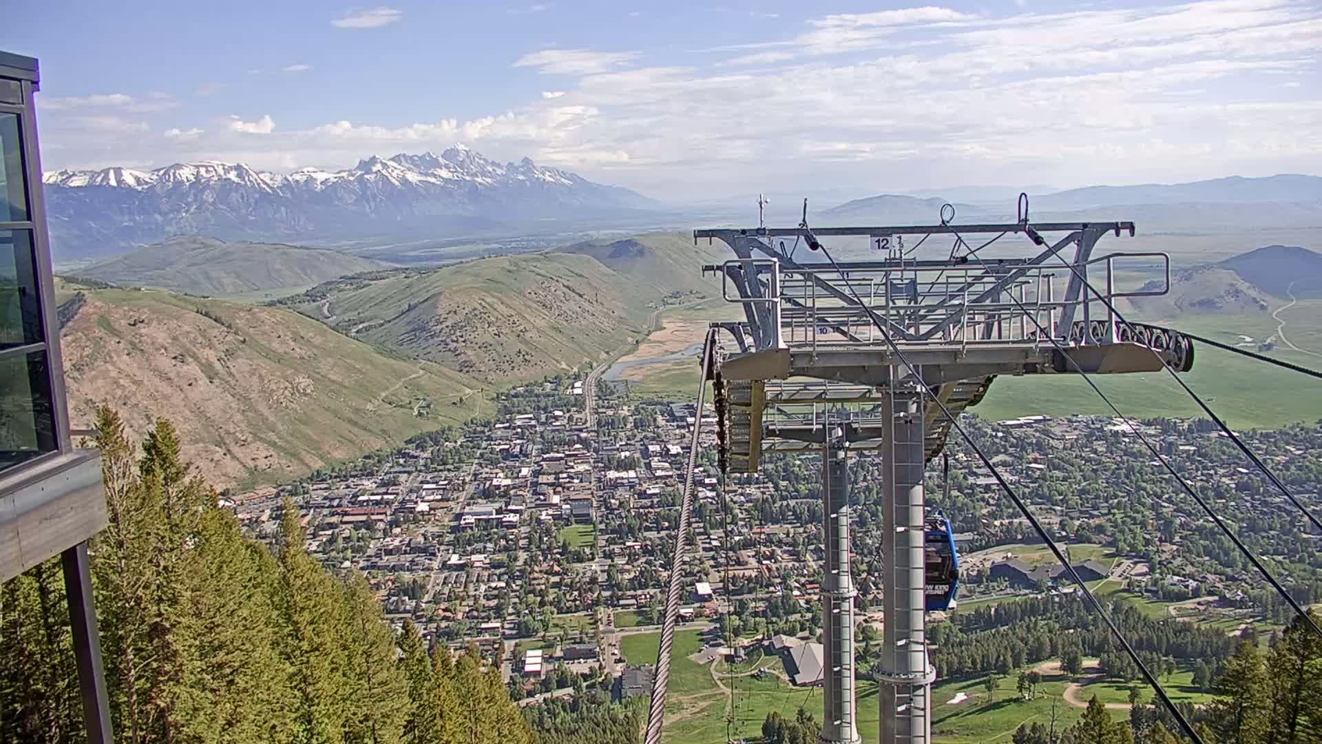 A chairlift overlooks a town nestled in a valley, with snow-capped mountains in the distance under a partly cloudy sky.