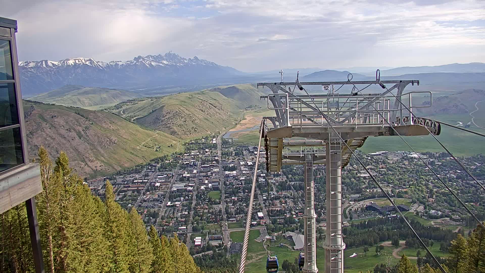 A chairlift ascends a mountain overlooking a valley town and snow-capped mountains under partly cloudy skies.