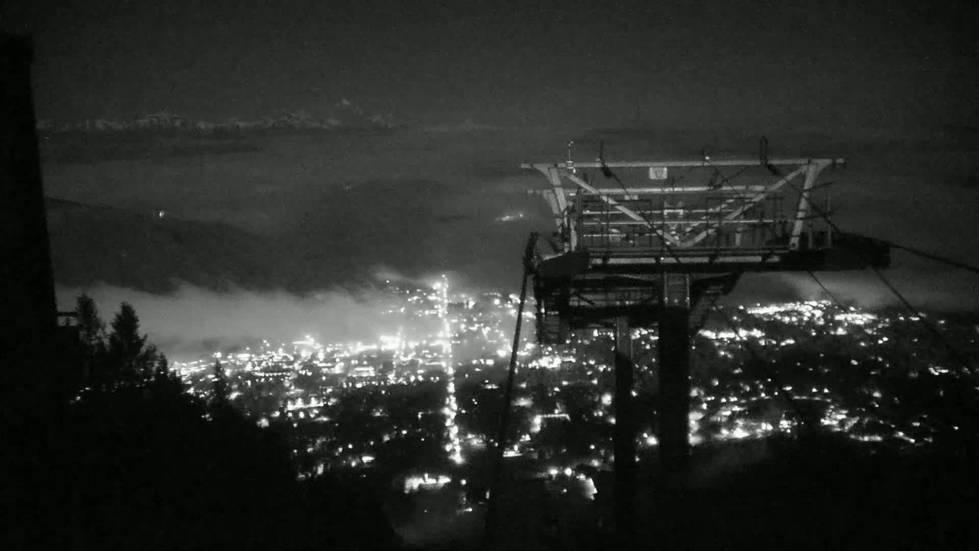 A nighttime view from a chairlift shows a valley town illuminated below, partially obscured by fog, with mountains visible in the far distance under a dark sky.