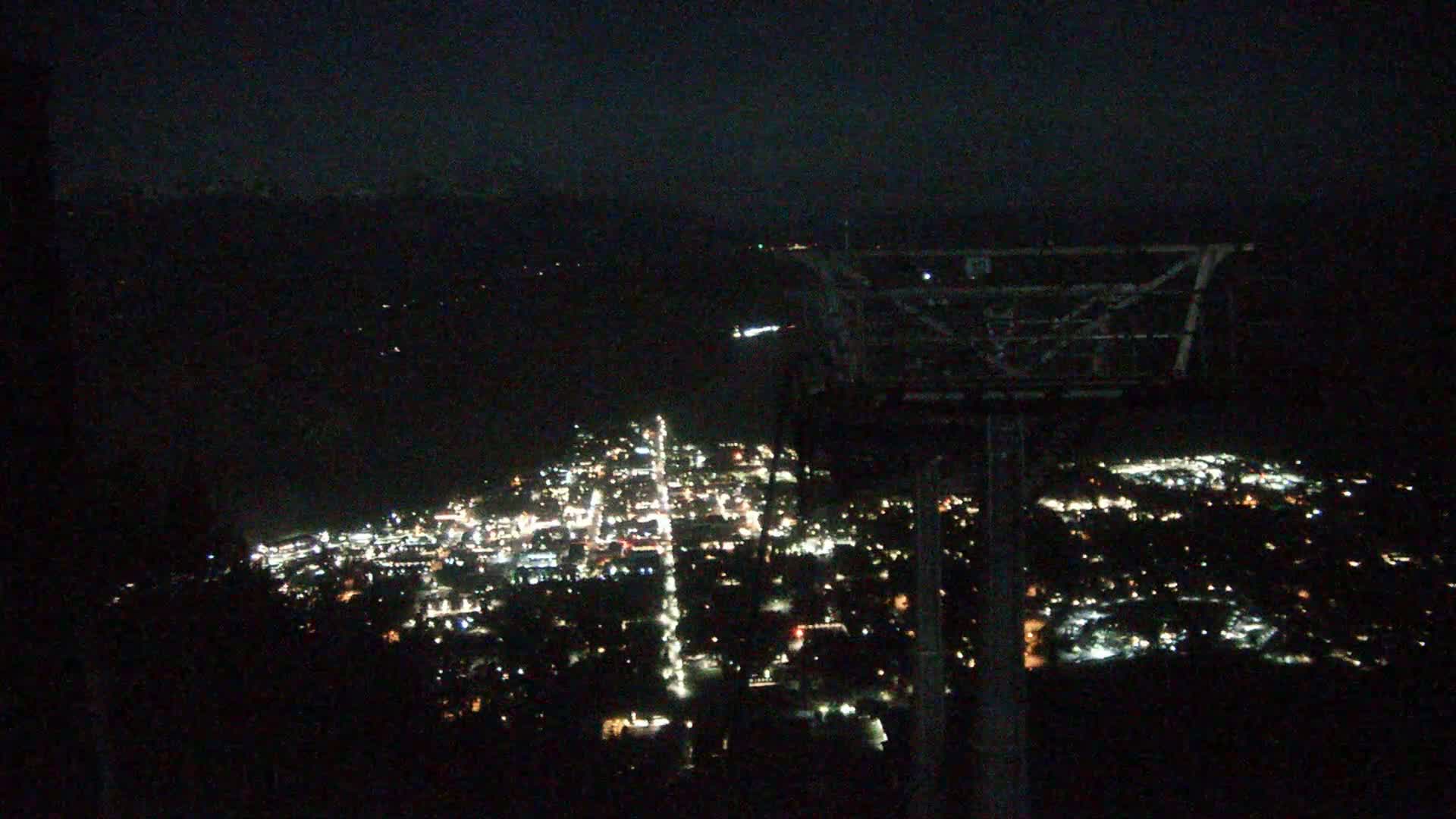 A nighttime aerial view shows a city illuminated with many small lights, viewed from a high vantage point next to a cable car structure, under a dark, clear sky.