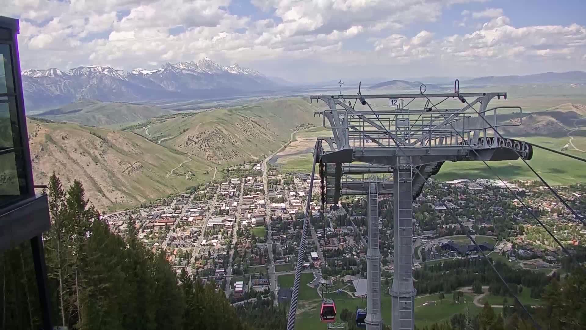 A chairlift ascends a mountain overlooking a town nestled in a valley, with snow-capped mountains in the distance under a partly cloudy sky.
