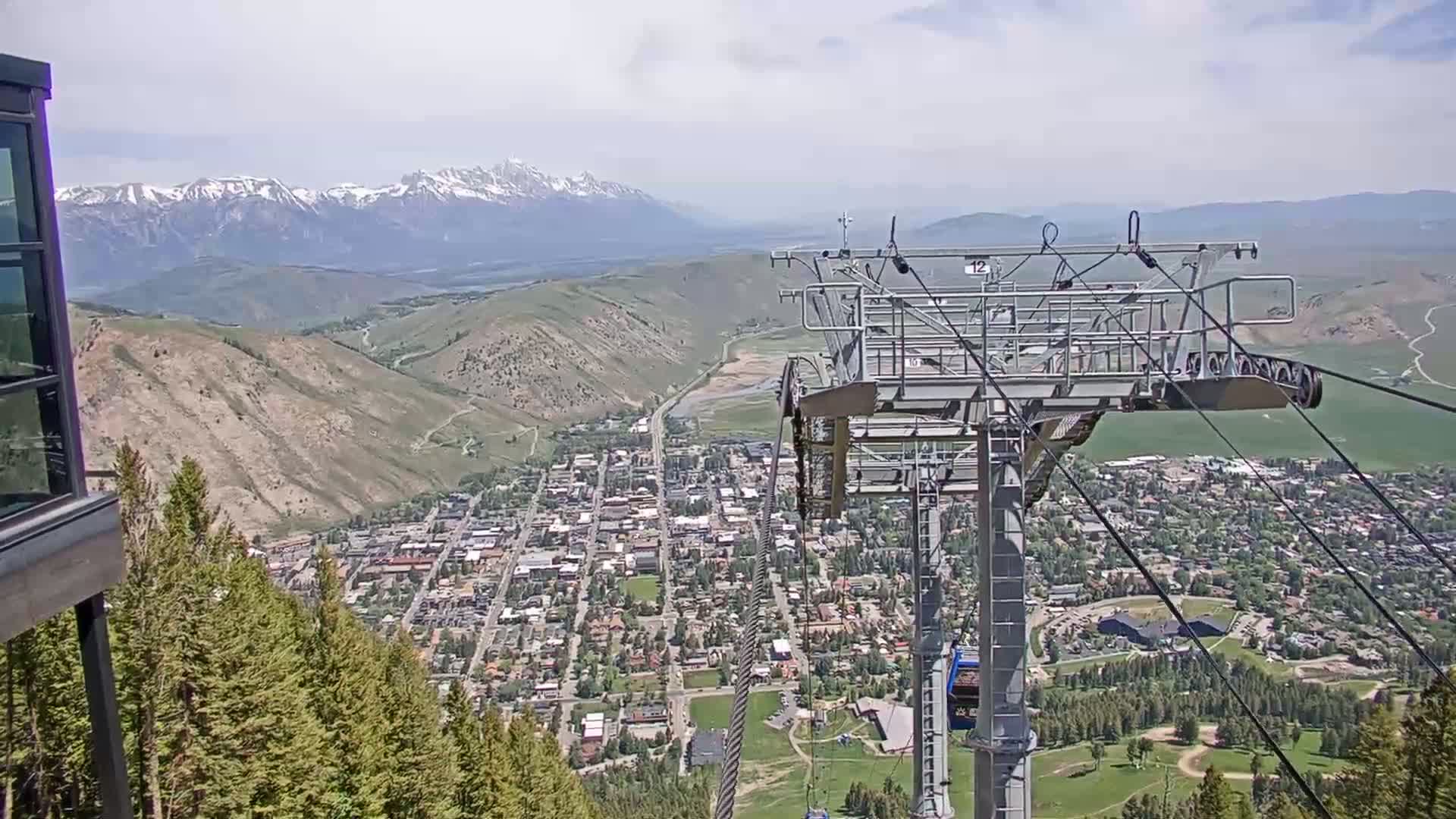 A chairlift ascends a mountain overlooking a town nestled in a valley, with snow-capped mountains visible in the distance under partly cloudy skies.