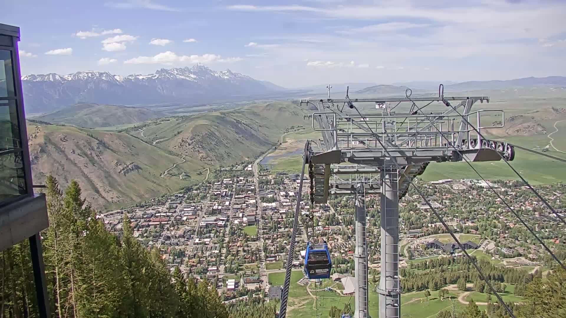 A chairlift ascends a mountain overlooking a town nestled in a valley, with snow-capped mountains visible in the distance under partly cloudy skies.